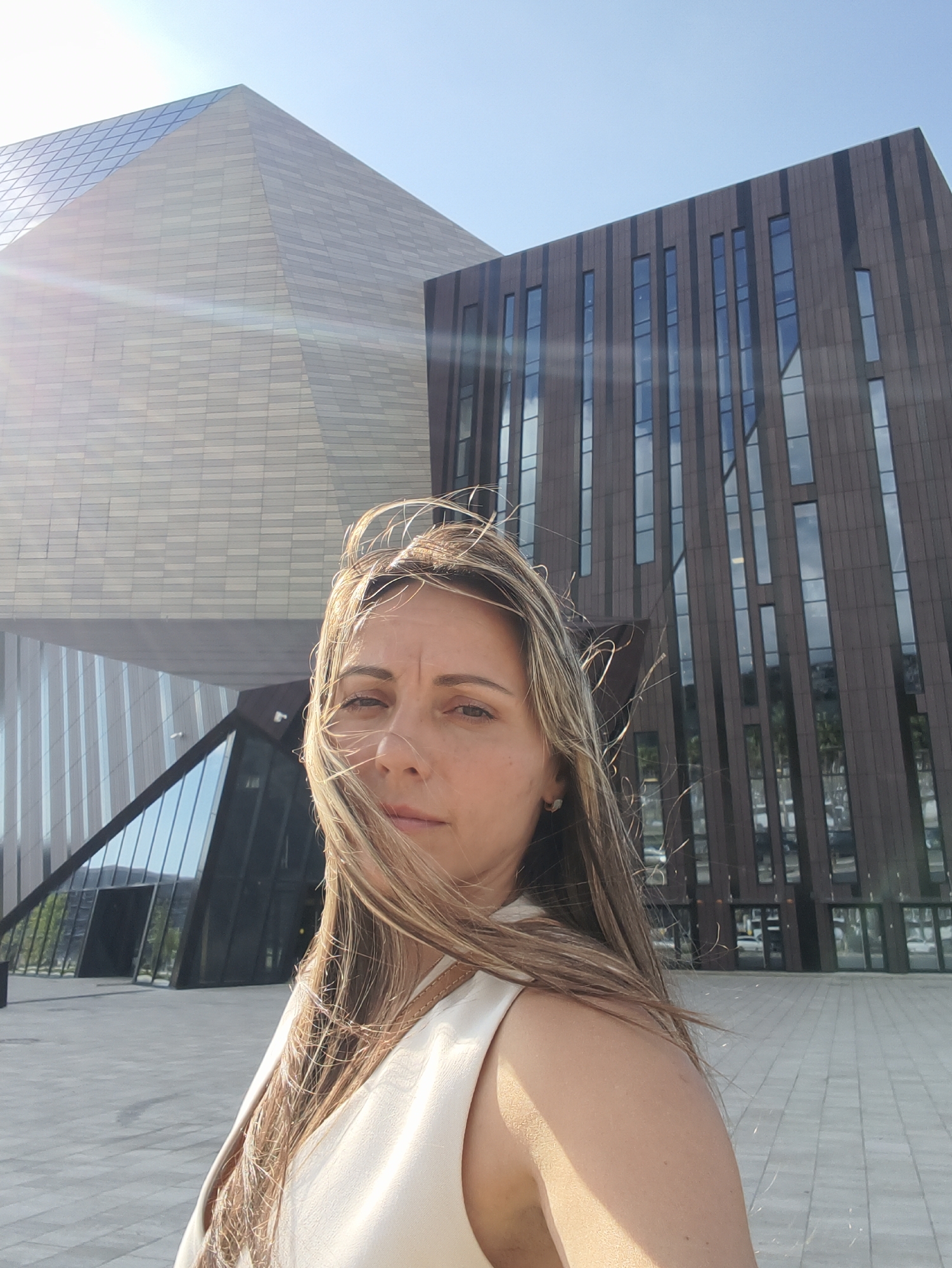 A woman with long, blonde hair is taking a selfie in front of the Sirius Federal Territory administrative buildings in Sochi, Russia. The woman is wearing a light-colored sleeveless top. Her hair is blowing in the wind. The buildings behind her are modern, with geometric shapes and large windows. The sky is clear and blue. The ground is paved with light-colored tiles. The sun is shining, creating lens flare in the upper left of the image.