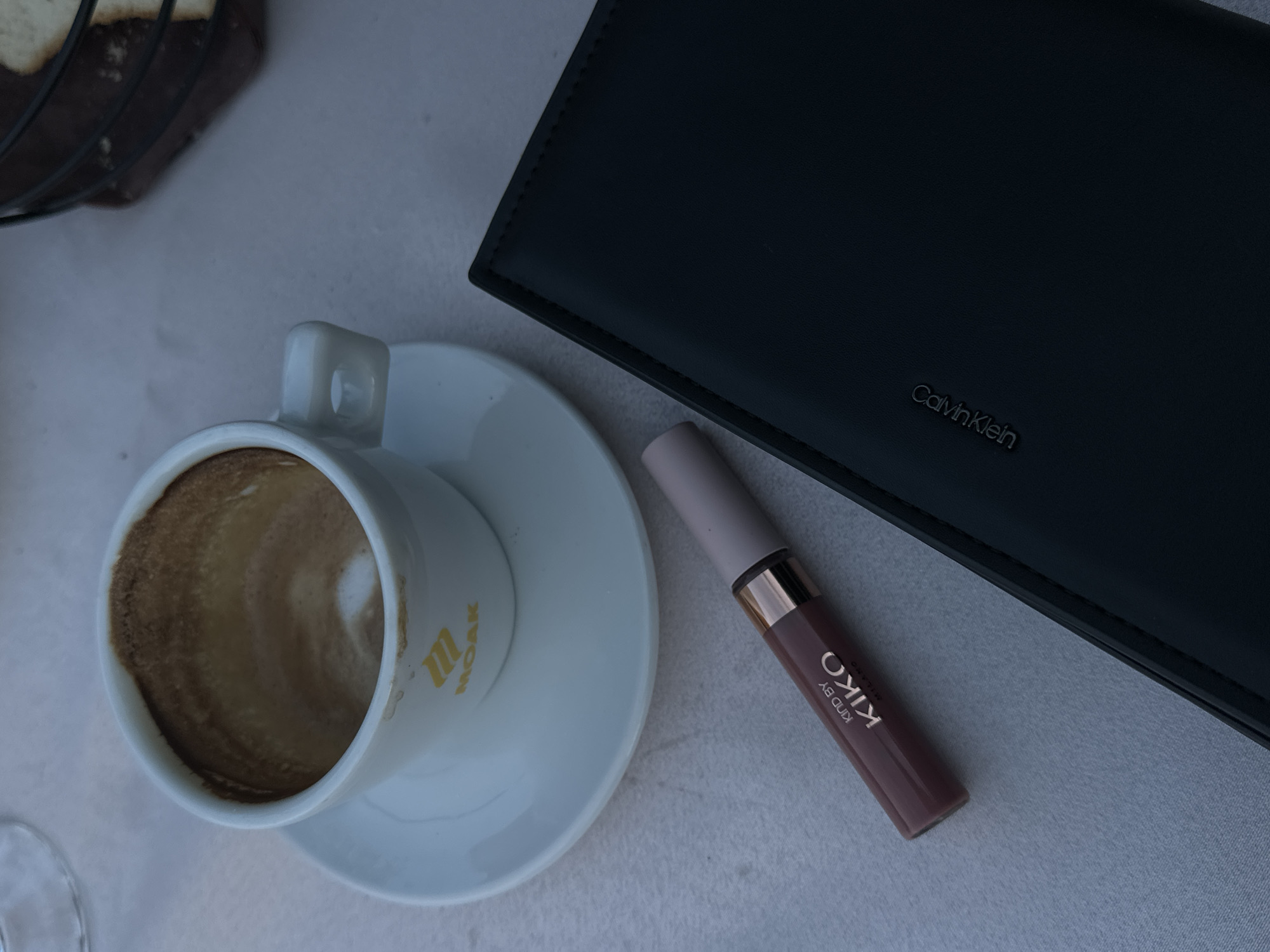 A close-up, overhead shot shows a white coffee cup with coffee residue on the inside and on the rim, resting on a white saucer. The cup has a logo that reads "MOAK" in yellow. To the left of the cup, a Kiko Milano liquid lipstick in a muted rose shade lies horizontally. To the right of the cup and lipstick is a black wallet or folder with the "Calvin Klein" logo embossed in silver. The background is a textured white surface, possibly a tablecloth. The scene is set in a cafe in Virpazar, Montenegro.