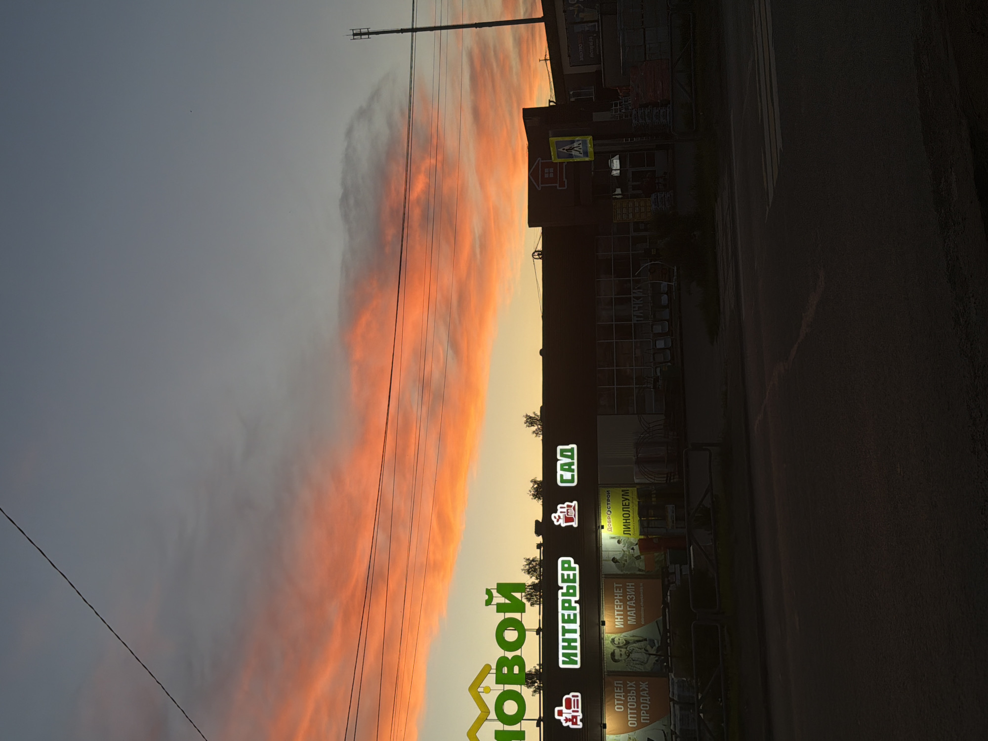 The image captures a dramatic sunset or sunrise over a commercial area in Chaikovsky, Russia. A striking, elongated band of vibrant orange and red clouds dominates the middle of the sky, contrasting with the muted blue-gray tones above. Below the colorful clouds, the horizon glows with a softer yellow-orange light.
On the right side, a large commercial building or complex is silhouetted against the sky. Its facade features several brightly illuminated signs with Cyrillic text. The most prominent sign displays "НОВОЙ" (NOVOY) vertically, and "ИНТЕРЬЕР" (INTERYER) and "САД" (SAD) horizontally, likely indicating a home goods or garden store. Below these, additional text reads "ИНТЕРНЕТ МАГАЗИН" (INTERNET MAGAZINE), "ОТДЕЛ ОПТОВЫХ ПРОДАЖ" (WHOLESALE DEPARTMENT), and "ЛИНОЛЕУМ" (LINOLEUM). A smaller yellow sign with a house icon and a pedestrian crossing symbol is also visible on the building.
Utility poles and power lines extend across the upper left and central parts of the sky. The lower right portion of the image shows a dark section, presumably a street or parking lot. There are no people or specific activities depicted. The scene conveys a tranquil, early morning or evening atmosphere, with the vivid natural light of the sky providing a striking backdrop to the urban commercial architecture.