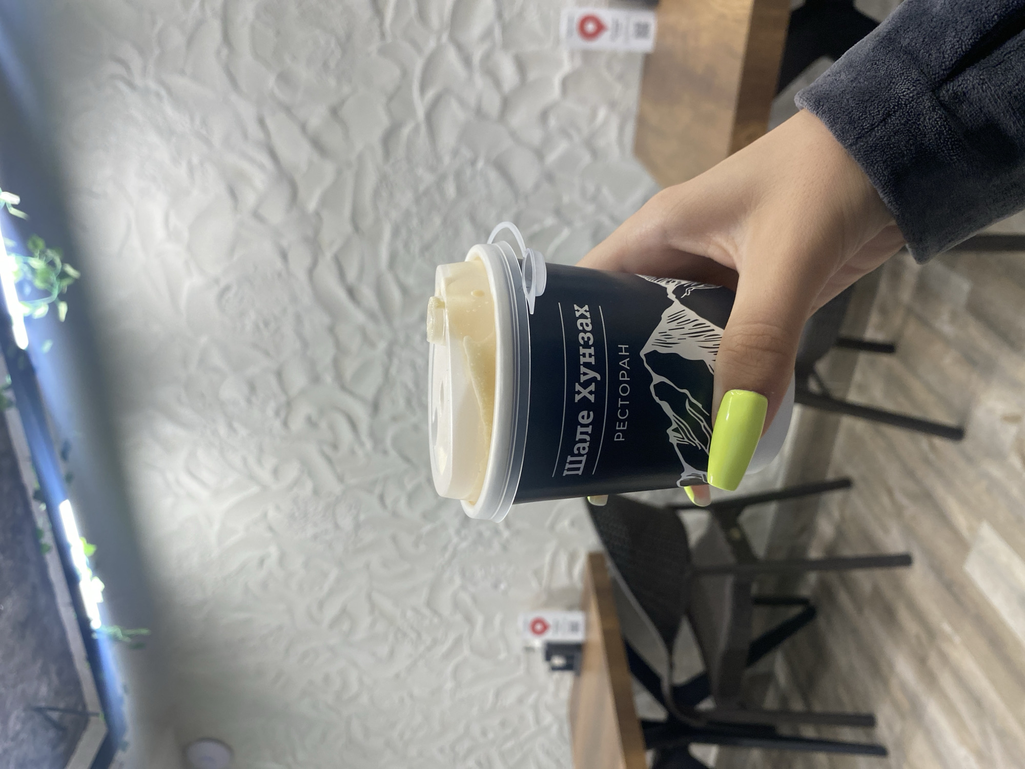 A person's hand, with bright neon green painted fingernails, holds a dark blue paper coffee cup with a white lid. The cup has white text that reads "Шале Хунзах" and "РЕСТОРАН" above a graphic of mountains. The background suggests an indoor setting, possibly a cafe or restaurant, with a textured white wall, tables with wooden tops and dark chairs, and overhead lighting with some artificial greenery. The overall impression is a casual moment captured inside an establishment.