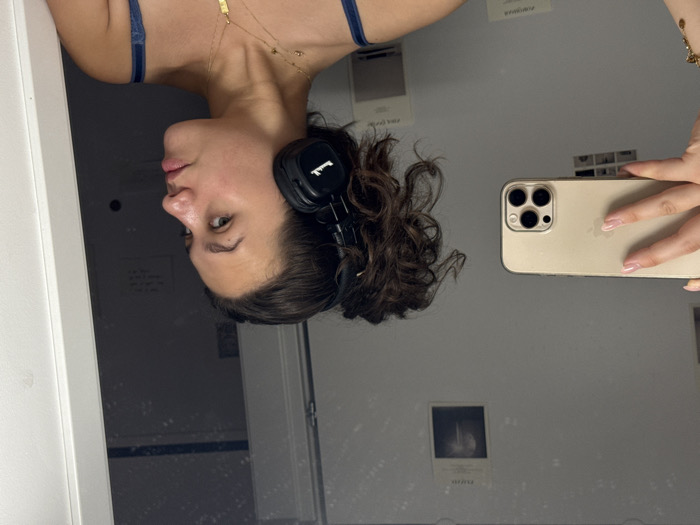 The image depicts a young adult female captured in a mirror selfie within an indoor setting. The specific location within Milan, Italy, cannot be visually confirmed as no identifying city landmarks or architectural features are present.
**Subject:** A female, visible from the upper chest to above the head, is the primary subject. She has dark, curly hair styled in an updo. Her skin tone is fair, and she appears to be wearing minimal or no makeup. Her gaze is directed to her right, away from the camera. She is wearing black Marshall-branded on-ear headphones on her left ear. Around her neck are multiple thin, gold-colored necklaces, one featuring a small rectangular pendant. Her garment appears to be a dark blue tank top or strap.
**Actions:** The individual is actively taking a self-portrait using a smartphone reflected in a mirror.
**Objects:** A gold-colored smartphone, consistent with an Apple iPhone due to the visible logo and triple-lens camera array, is held in her left hand, which has light-colored manicured nails. Two small, rectangular items, possibly photographs or notes, are taped to the light-colored wall reflected in the mirror behind the subject.
**Environment:** The background consists of a plain, light-colored wall (likely white or off-white) visible through the mirror's reflection. A vertical white panel or door frame is visible on the far right side of the frame. The reflection within the mirror also reveals a dark opening or doorway behind the subject, suggesting an adjacent room or hallway. The scene indicates an interior, possibly domestic or private, space.