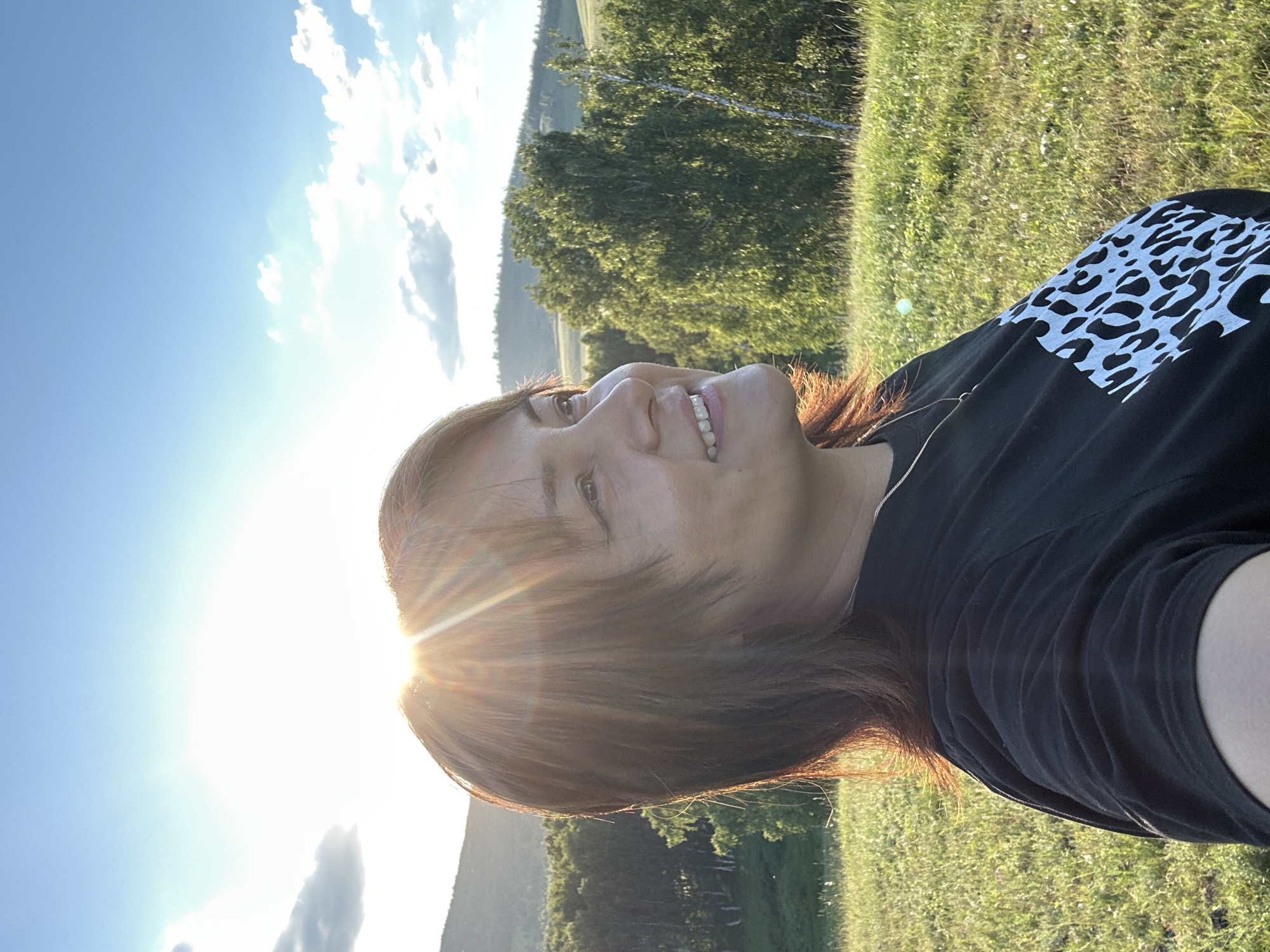 A woman with brown hair is captured from the chest up in a portrait taken outdoors. She is looking to her right, with a slight smile, her face illuminated by the setting sun which creates a lens flare effect. She is wearing a black t-shirt with a white leopard print design on the front. The background consists of a lush green grassy field, a line of trees in the middle ground, and rolling hills in the distance under a bright blue sky with scattered clouds. The overall lighting suggests it is late afternoon or early evening.