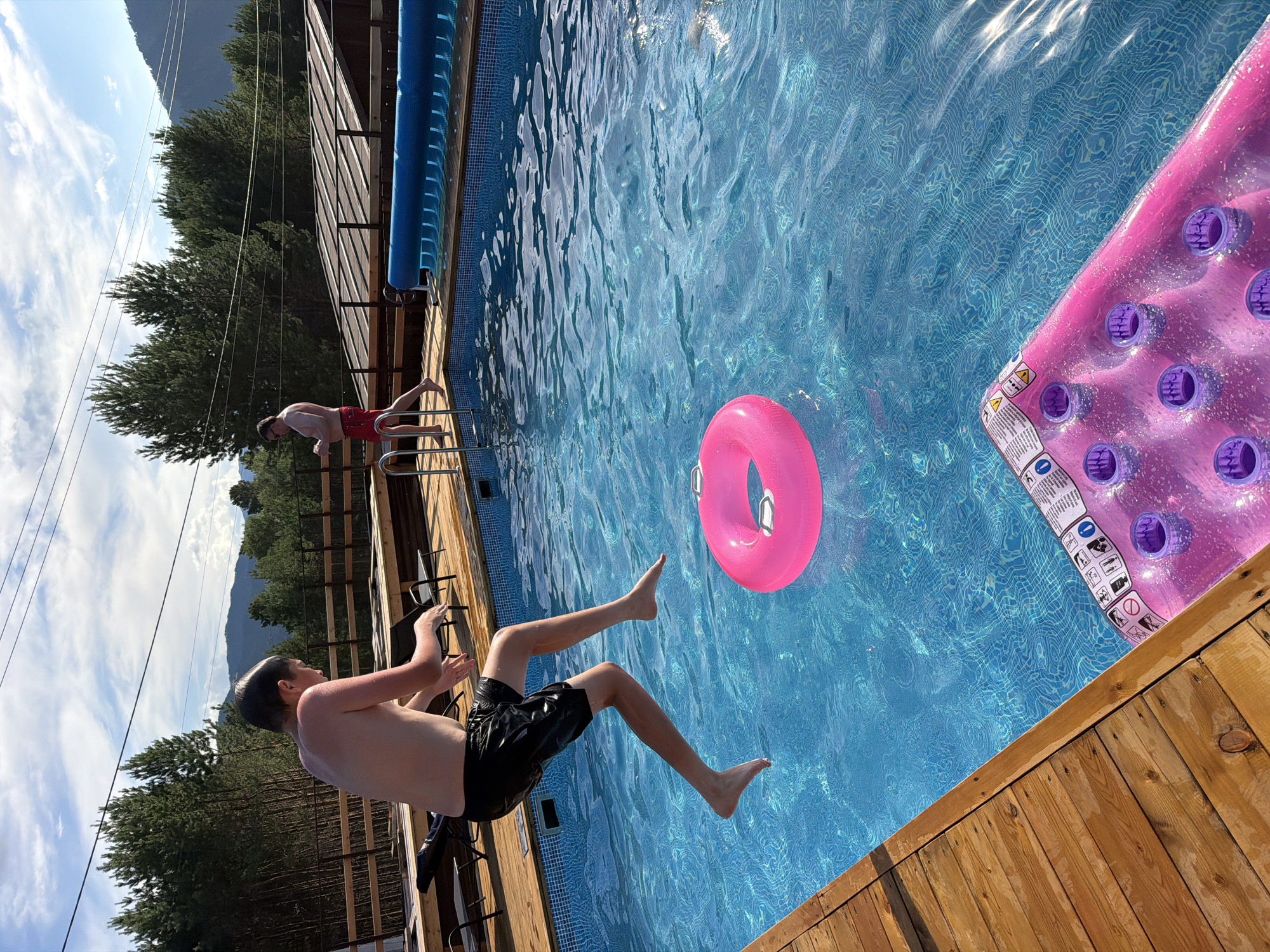 A boy is captured mid-air as he jumps into a bright blue swimming pool on a sunny day in Elekmonar, Russia. The pool is surrounded by a wooden deck, and in the distance, pine trees and mountains form a scenic backdrop. A pink inflatable ring floats in the water, and a pink inflatable mattress with cup holders rests at the edge of the pool. Another man in red shorts stands near the edge of the pool, looking at his phone. The scene conveys a sense of summer recreation and outdoor enjoyment.