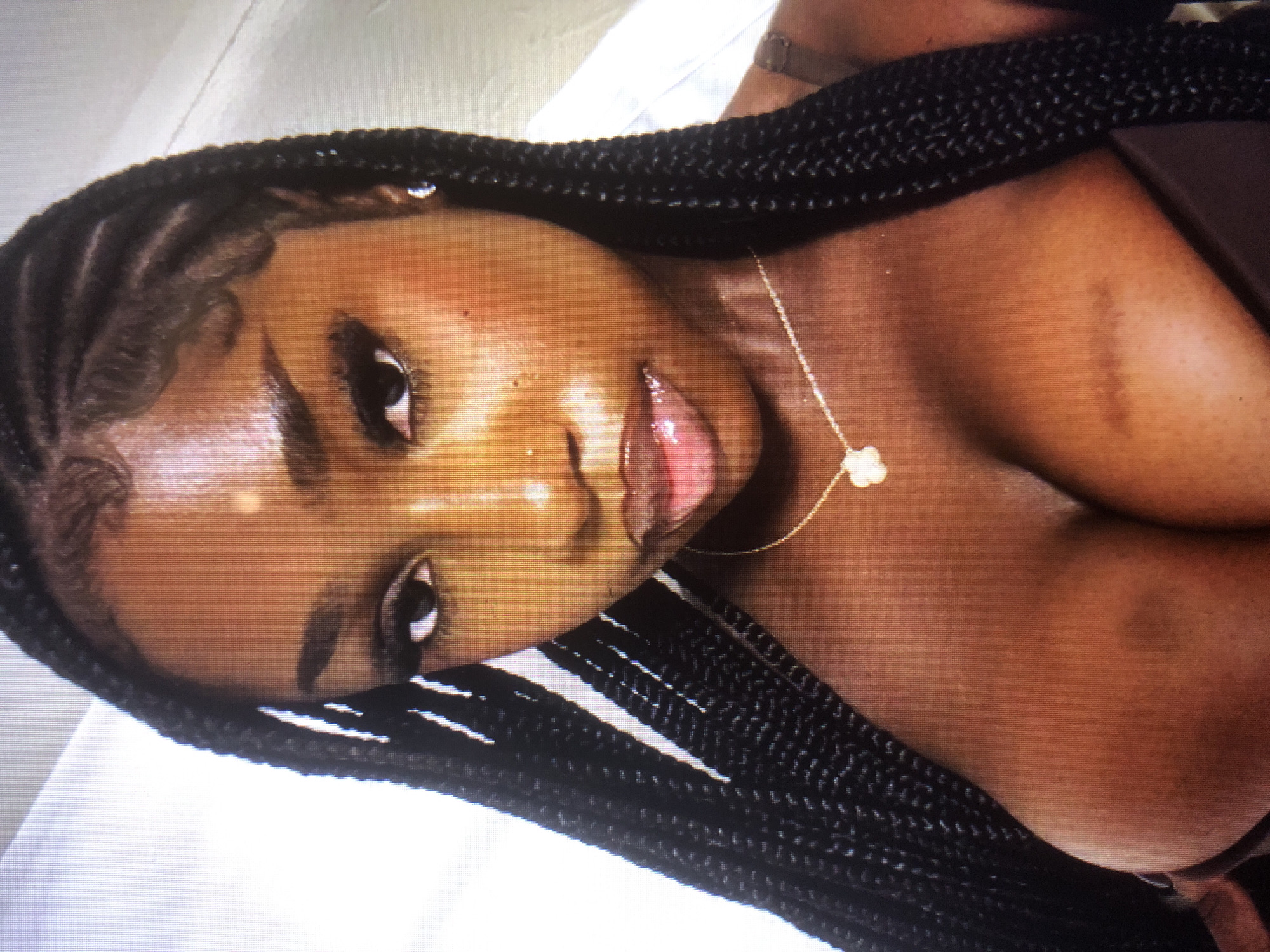 The image is a close-up, chest-up selfie of a young Black woman with dark braided hair. She is wearing a brown top and a delicate gold necklace with a clover-shaped pendant. Her skin is a rich brown tone, and she has a small nose stud. The lighting highlights her dewy complexion and glossy lips. Her braided hair is styled with baby hairs around her hairline. The background is largely out of focus, appearing to be a neutral, indoor setting. There are no discernible details that would place the image in Barakin Lerawi, Nigeria.