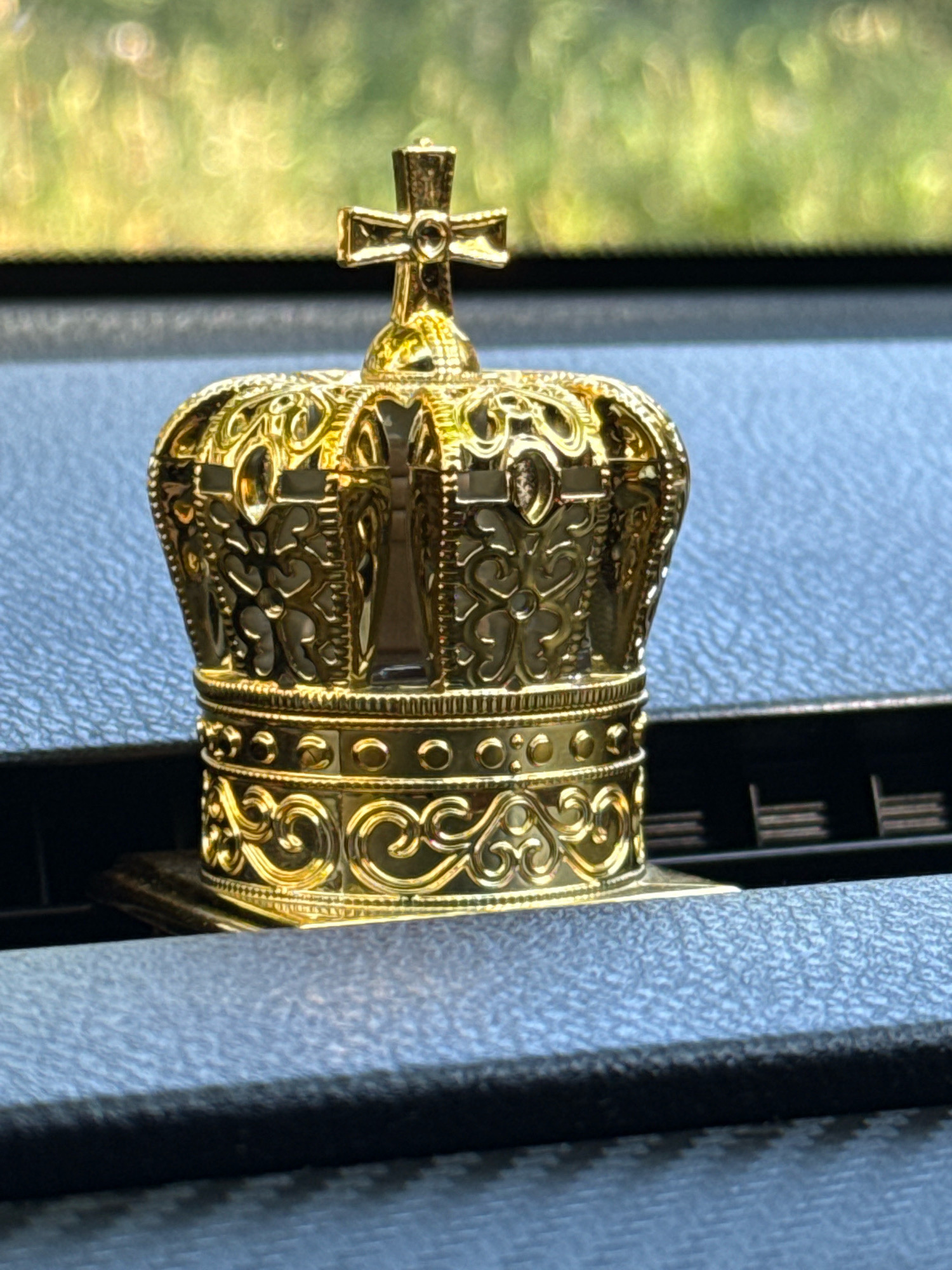 A close-up, eye-level, medium shot captures a golden crown-shaped car air freshener. The object is positioned on a dark gray textured dashboard. The crown features intricate filigree designs and a small cross on top. The background is blurred, showing a bright green and yellow bokeh effect, suggesting outdoor foliage.