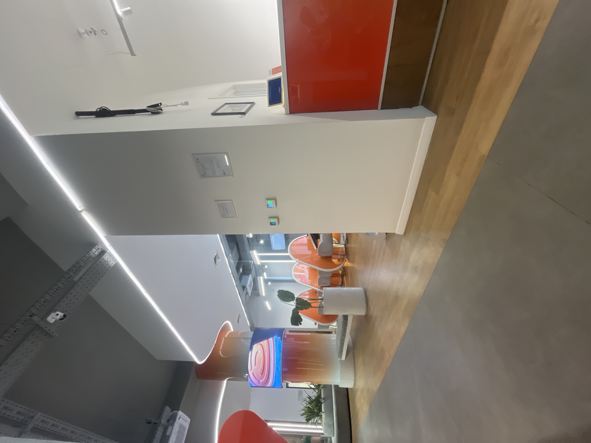 The image depicts the interior of what appears to be a modern office or commercial space in Yerevan, Armenia. The color scheme is predominantly white, orange, and brown. On the ceiling, there are visible lighting fixtures and a cable management system. A white wall with an orange panel and a digital display can be seen. There are certificates and other small objects attached to the wall. Orange chairs and tables are arranged in a waiting area. A plant is placed in a white pot. There are also monitors on poles with colored lights on the front of them. The floor is a mix of wooden and concrete surfaces. The image is taken from a slightly rotated angle.