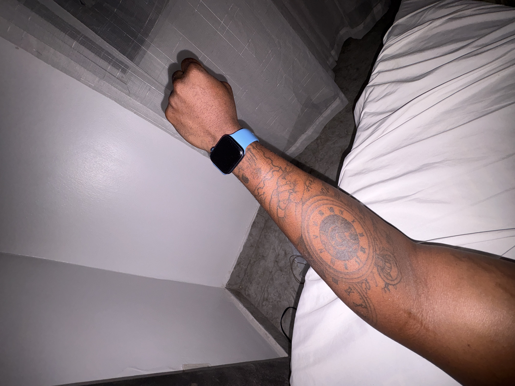 A tanned arm, tattooed with a clock face and other designs, wearing a blue smart watch, lies on a white sheet. The arm appears to be resting near a wall and a window with a semi-transparent curtain. The location is unknown; Iddo Tudunwada, Nigeria, cannot be determined from the provided image.