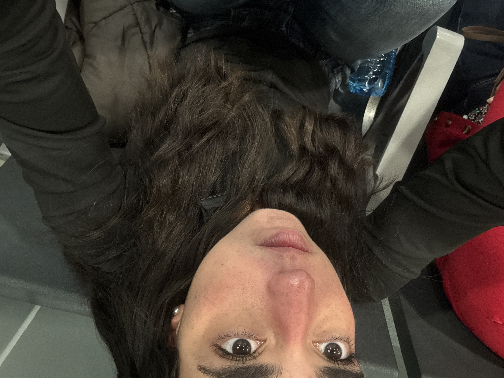 The image features a person, likely female, captured from an inverted low-angle perspective. Her head and upper torso fill a significant portion of the frame, with her dark, long, wavy hair fanning out towards the top of the image. She is wearing a dark, possibly black, long-sleeved jacket or top. Her face is visible from the nose downwards, with wide, dark eyes looking directly at the camera. She has distinct dark eyebrows and eyelashes, and scattered freckles are visible across her nose and cheeks. Notable dark smudges or markings, possibly eye black or smudged makeup, are present under her left eye. A small silver stud earring adorns her left ear.
The background suggests an indoor public setting. Elements visible include denim fabric (likely jeans) and a blue plastic water bottle towards the upper right. A bright red bag or piece of clothing is partially visible in the bottom right. A light grey, metallic-looking bar, possibly part of a public bench or seating, diagonally traverses the mid-ground. The overall scene implies a waiting area, such as an airport, train station, or similar public transport hub.
The activity appears to be the taking of a playful or unconventional selfie, indicated by the upside-down orientation and the subject's wide-eyed, engaging expression. No visible text is present in the image. The time of day and weather are not discernible due to the indoor setting.