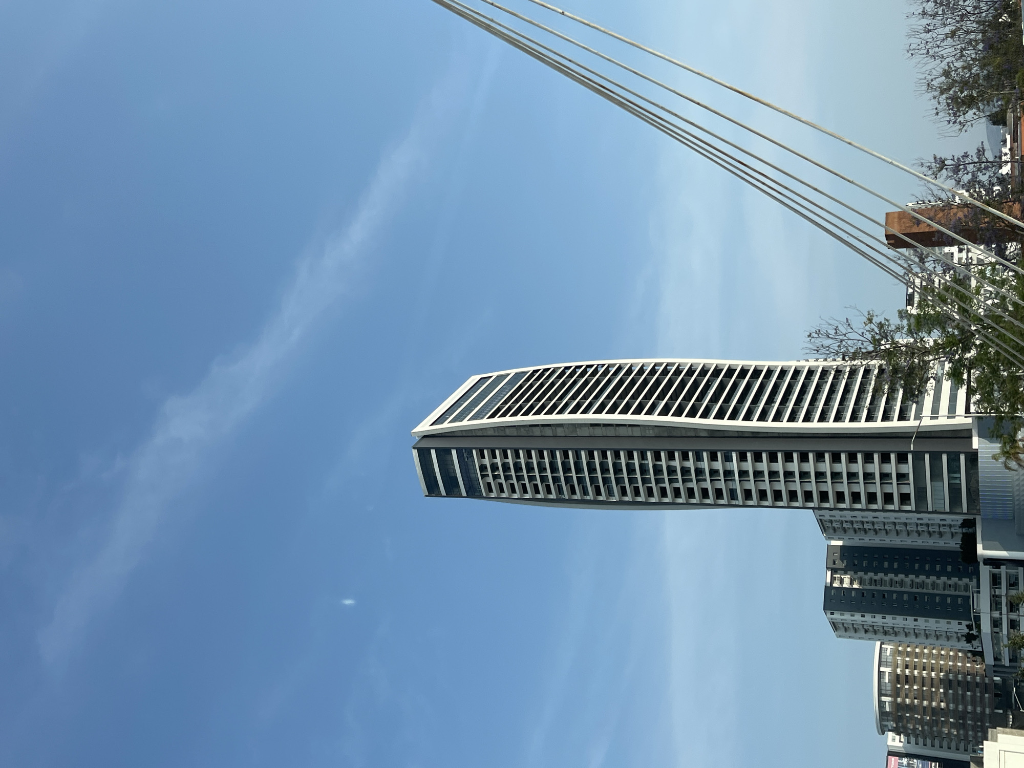 The image displays a modern urban scene in San Bernardino Tlaxcalancingo, Mexico, under a bright blue sky with faint, wispy clouds. The central focus is a tall, contemporary high-rise building on the left, characterized by its distinctive curved facade and a series of prominent white horizontal fins or louvers on its upper stories. Below these fins, the building features a grid pattern of windows and darker panels.
In the foreground, running diagonally across the upper right portion of the frame, are several parallel cables or wires, suggesting proximity to a bridge or another architectural structure. To the right of the main skyscraper, and partially visible through the cables and some trees with green foliage and hints of purple flowers, are other multi-story buildings. These structures exhibit modern architectural styles, with some featuring dark gray and white paneling, contributing to the urban density of the scene.
The photograph captures a static, daytime view with clear lighting, indicating fair weather. There are no people visible, nor are there any discernible activities taking place. No legible text appears in the image. The overall impression is one of contemporary architecture set against an open sky in a developing urban landscape.