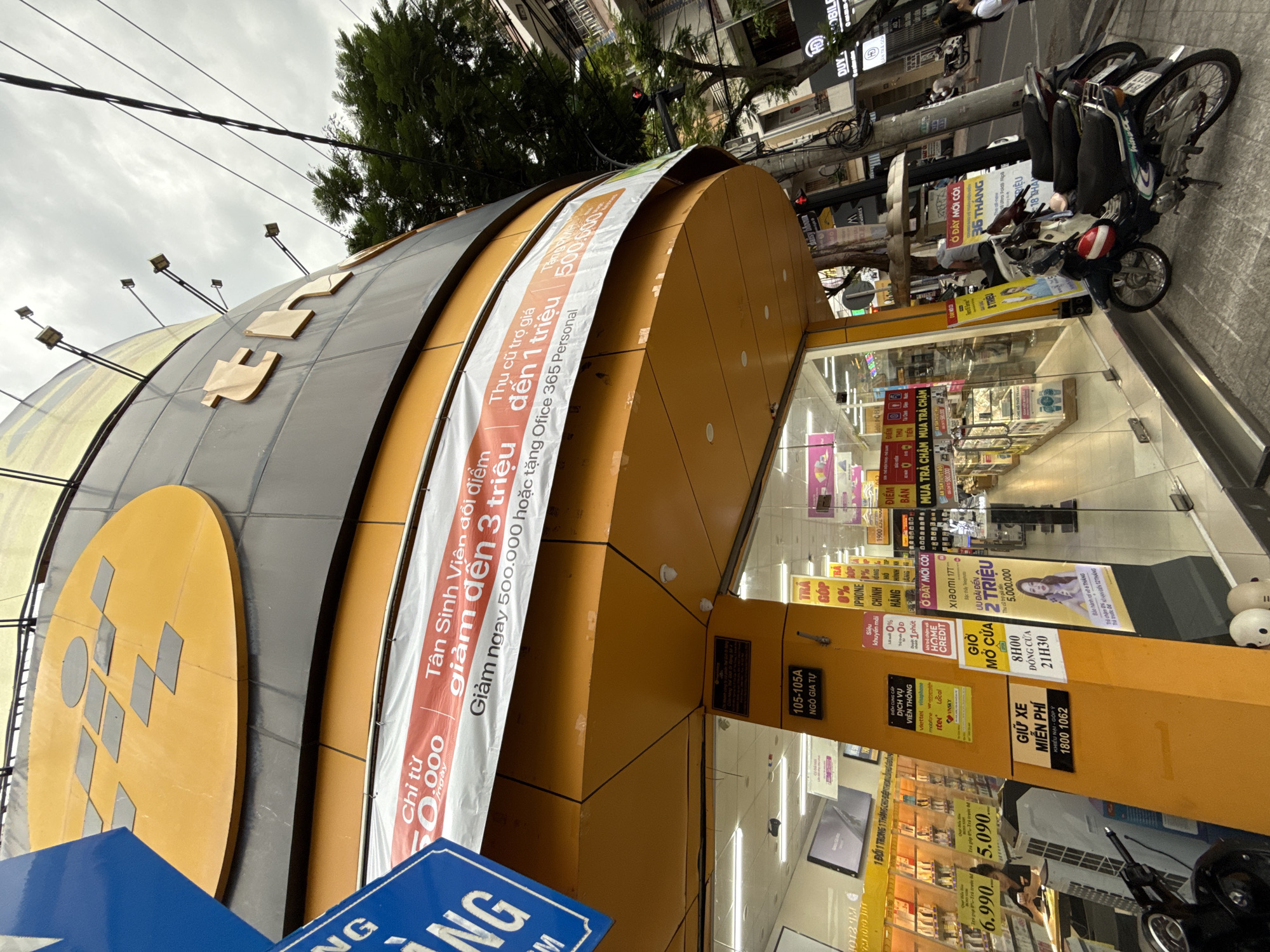 The image shows a street view of a "The Gioi Di Dong" electronics store in Nha Trang, Vietnam. The store's exterior features a prominent yellow and orange facade with large signage. A blue sign with white text is visible on the left side of the image. Banners on the building advertise discounts and promotions, including "Giảm ngay 500.000 hoặc tặng Office 365 Personal" and "Tân Sinh Viên đổi điểm giảm đến 3 triệu".
Inside the store, displays of electronic devices, such as phones and watches, are visible through the glass storefront. Several motorcycles are parked on the sidewalk in front of the store. A sign near the entrance indicates parking is free for motorcycles. People are seen walking on the sidewalk and potentially inside the store. The overall environment suggests a commercial street in a Vietnamese city, with utility wires and trees lining the street.