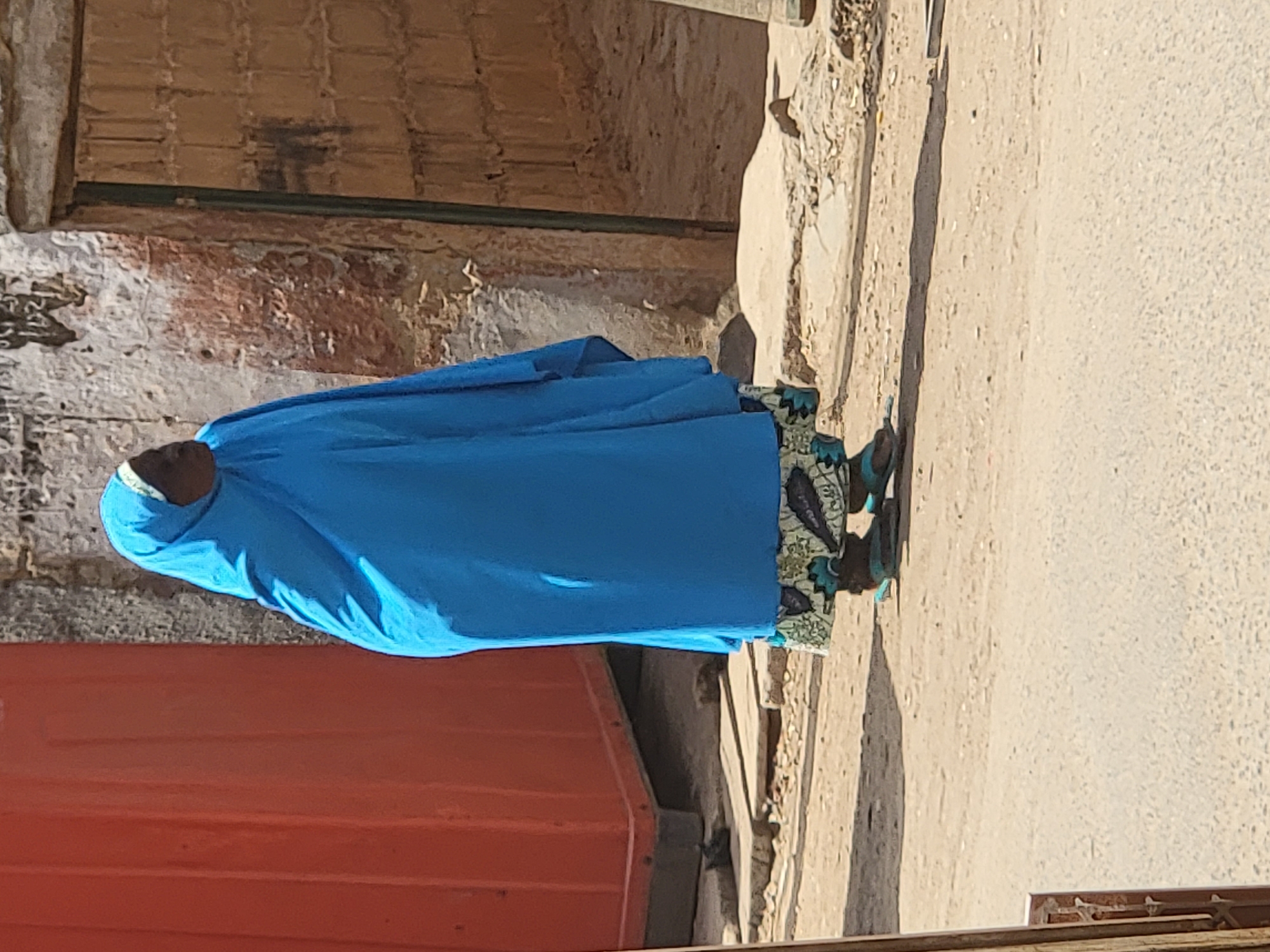 This factual description is based on the provided image and its crops, set in Maiburiji, Nigeria.
The image features a person, likely a woman, standing on what appears to be an unpaved street or pathway. The individual is largely seen from the side and slightly from behind, wearing a bright, full-length, flowing blue garment that covers the body and head. The lower edge of this garment features a distinct patterned border in shades of green, blue, and black motifs. The person is wearing dark shoes, possibly black, with small greenish-blue details.
To the left of the subject, occupying a significant portion of the lower-left foreground, is a large, reddish-orange object with prominent horizontal ridges, which could be part of a container, a low structure, or a vehicle. Behind the person, there's an aged and weathered wall constructed from bricks or rough stone, displaying patches of lighter and reddish-brown coloration. A horizontal dark green bar or pipe runs along the upper part of this wall.
The ground to the right of the person is an unpaved, dusty surface, suggesting a street or path in a rural or semi-urban setting. Strong, well-defined shadows are cast on this surface by the person and other unseen objects, indicating bright sunlight and clear weather, likely around midday or early afternoon. No specific activity is clearly depicted beyond the person's presence in the scene. There is no visible text within the image, and due to the garment, the person's facial expression or emotions are not discernible.