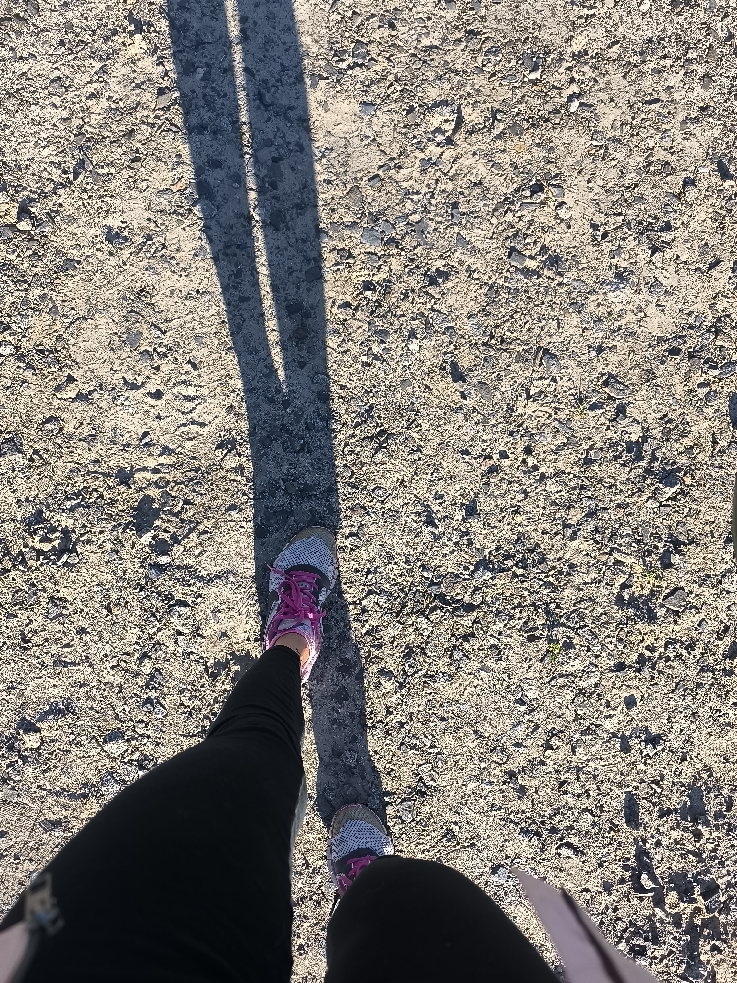 The image depicts a person's legs and feet wearing black leggings and athletic shoes with pink laces, standing on a gravelly path. A long shadow is cast by the person, indicating sunlight. The environment is an outdoor, unpaved area. The location in Horishni Plavni, Ukraine, is not identifiable from the visual information provided in the image.