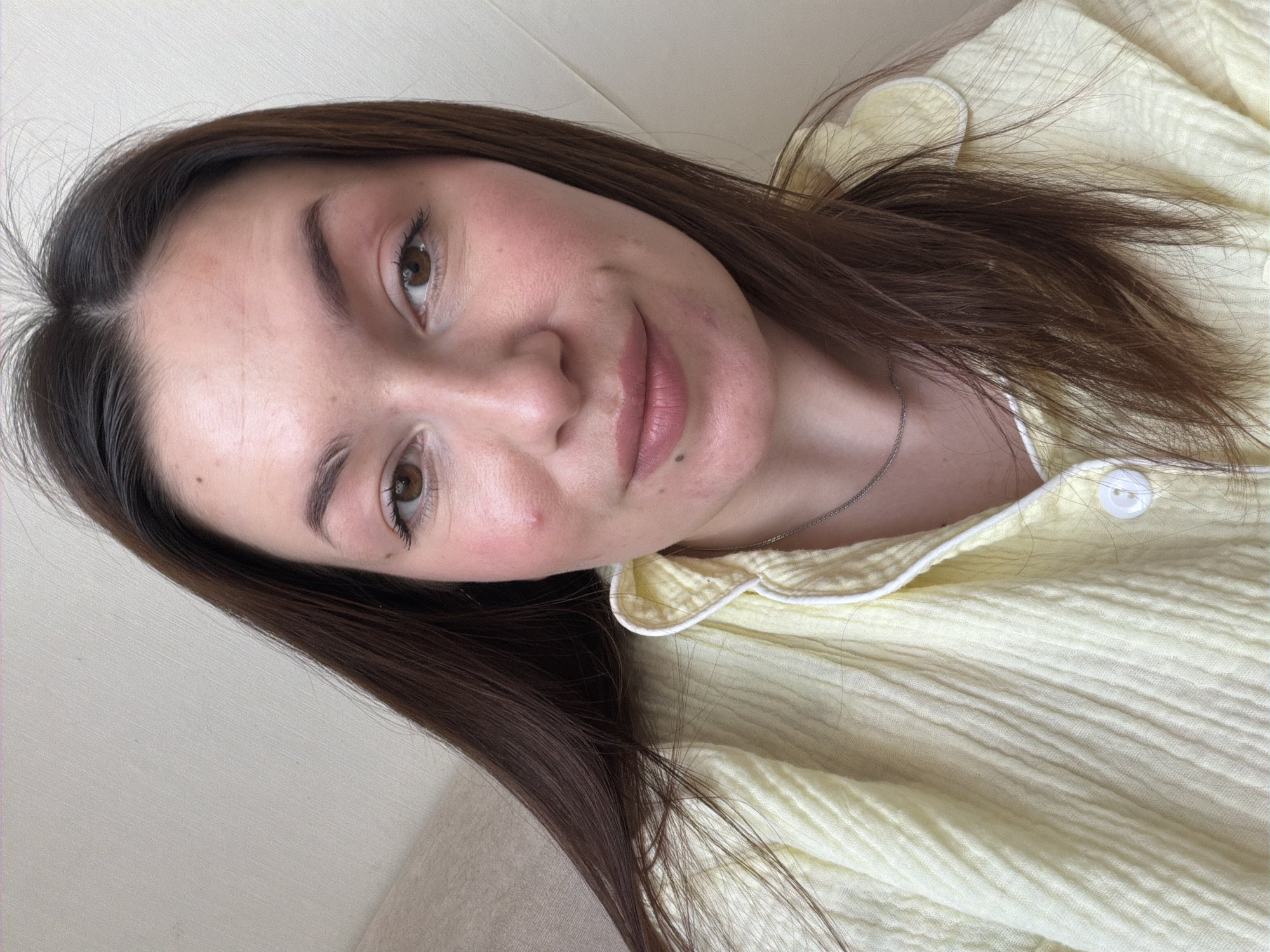 A woman is centered in the image, looking at the camera. She has light skin, brown hair, and brown eyes. Her facial expression is neutral. She is wearing a pale yellow top with white trim around the collar. A thin silver necklace is visible around her neck. The background appears to be a light beige wall or surface. There is no information in the image to confirm a location in the city of Izhevsk, Russia.