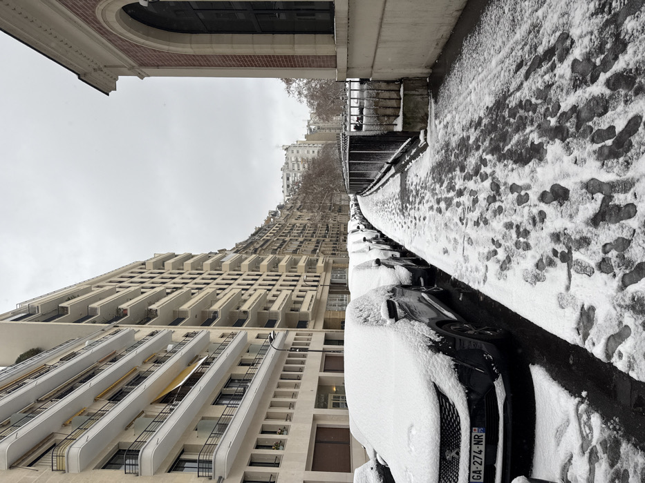 The scene depicts a snow-covered urban street in Paris, France, under an overcast grey sky. No individuals are visible. On the left, a row of multi-story residential buildings, primarily light-colored stone or stucco with prominent balconies featuring black metal railings, extends into the distance. A streetlamp is positioned in front of these buildings. Below, a line of parked automobiles, each covered by a layer of fresh snow, occupies the street. The closest vehicle is a dark-colored Peugeot with a visible French license plate "GA-274-NR".
To the right, a snow-covered sidewalk stretches into the background, marked by numerous discernible footprints indicating recent pedestrian movement. Adjacent to the sidewalk, a dark metal fence separates the path from a lower-lying area, possibly a park or garden, where bare trees are visible. Further right, a building with reddish brickwork and light-colored stone trim features a large, dark, arched window. The ground surface shows a mixture of settled snow and areas where the asphalt is exposed, particularly in the tire tracks of the vehicles. The overall environment suggests a cold, winter day with recent snowfall.