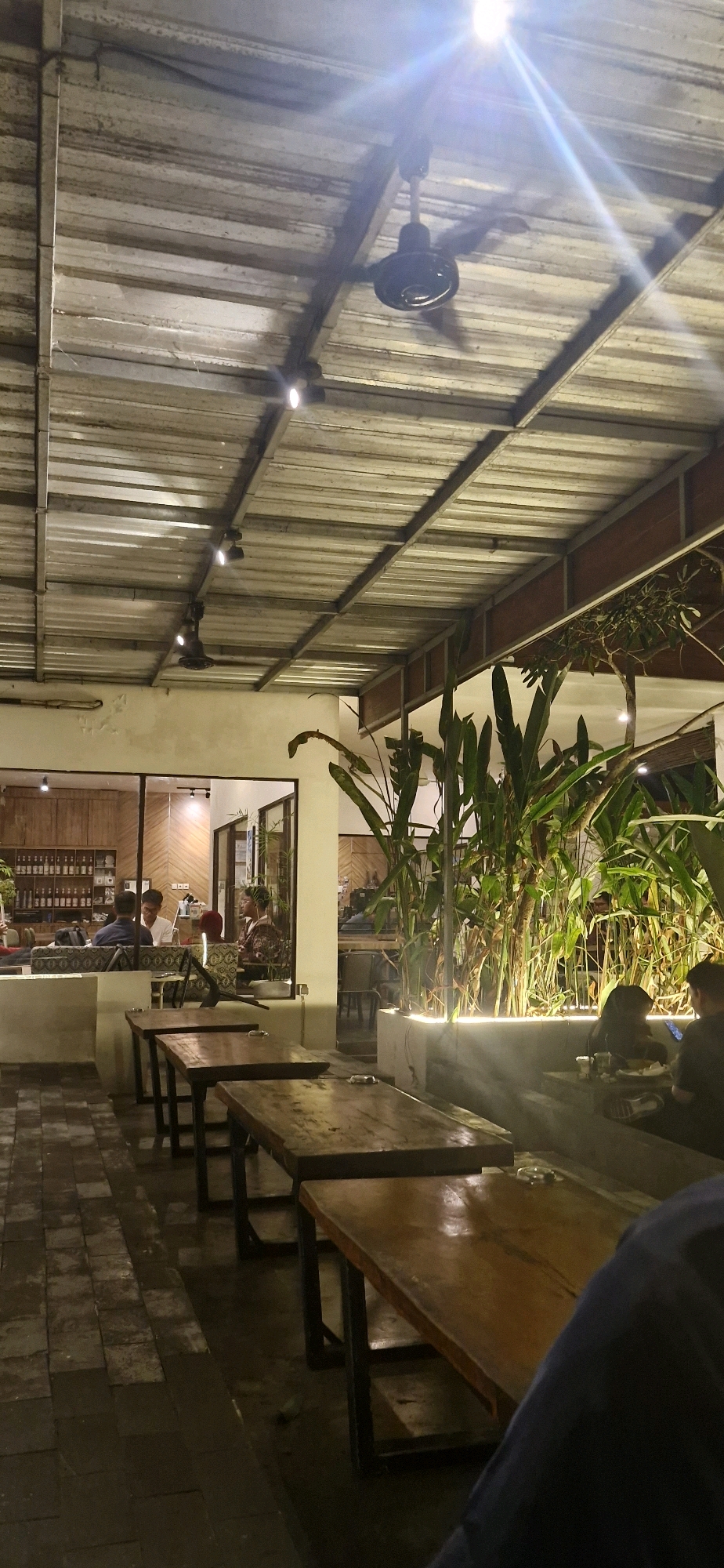 This image depicts the interior of a cafe in Banjarbaru, Indonesia, with a focus on the outdoor seating area. The scene is illuminated by warm lighting, highlighting the wooden tables and chairs arranged on a tiled floor. In the background, through large glass windows, glimpses of the cafe's interior are visible, showing customers seated at tables and a bar area stocked with bottles. Lush green plants are strategically placed, some in planters with integrated lighting, adding a touch of nature to the space. A ceiling fan is visible, suggesting a warm climate. The overall atmosphere appears relaxed and inviting, suitable for a casual gathering or a quiet moment.