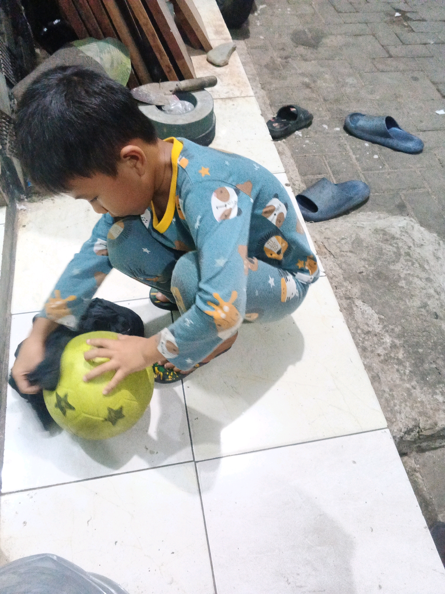 A young boy, likely Indonesian, is crouched on a tiled surface, engaged in wiping a yellow ball with black star patterns. He is wearing blue pajamas with cartoon animal prints and yellow trim. Several pairs of dark-colored flip-flops are scattered on the adjacent paved ground. The environment appears to be an outdoor area in the city of Kadu, Indonesia, characterized by a mixture of tiled flooring and rough concrete pavement. In the background, there are wooden planks, a metal fan, and a mortar and pestle. The lighting suggests it is either dusk or an overcast day.