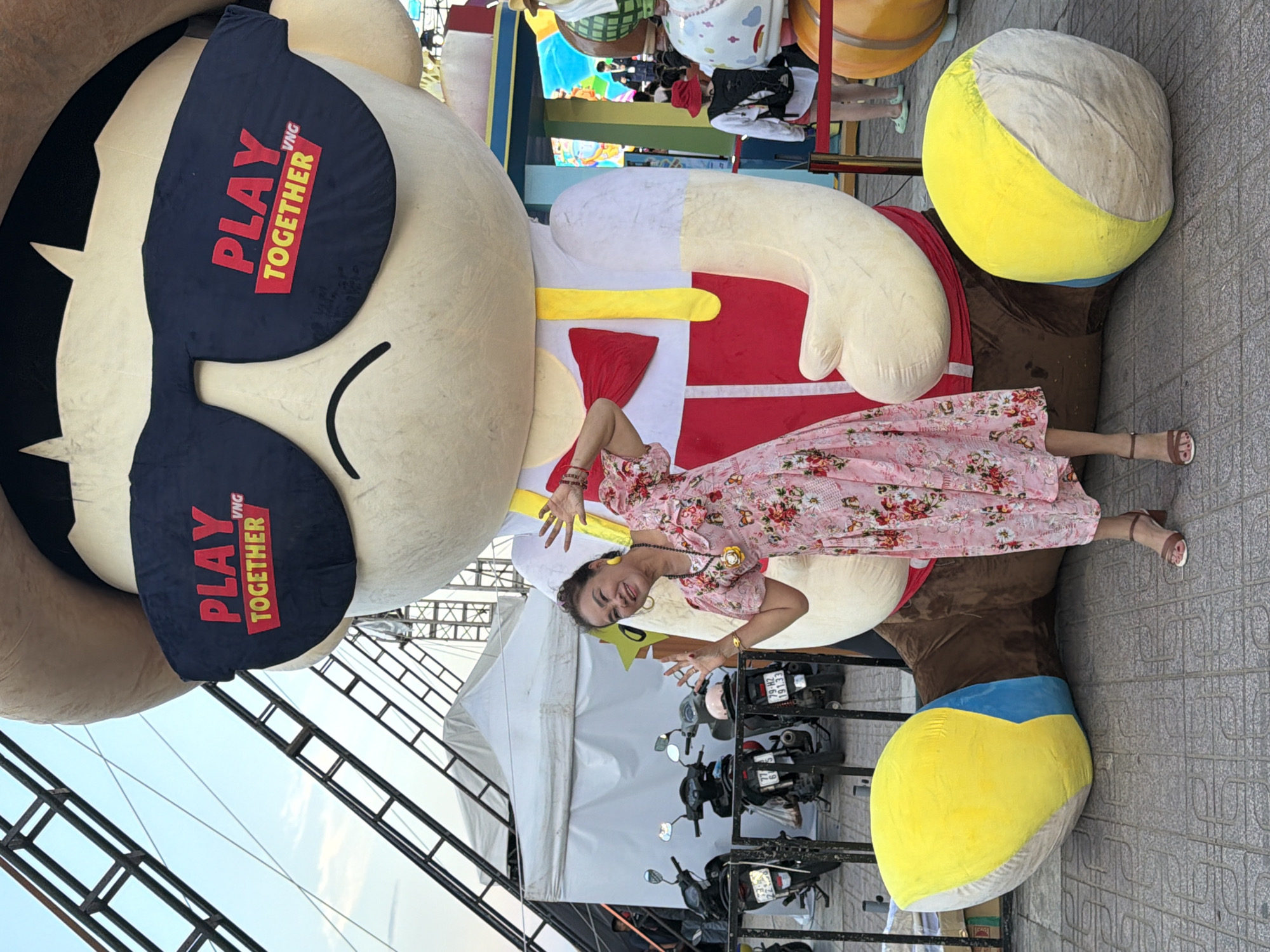A woman in a pink floral dress and brown heels poses in front of a large inflatable character with sunglasses that say "PLAY TOGETHER VNG." The inflatable character is wearing a red and white outfit with a yellow bow tie. In the background, there are tents, motorcycles, and people. The scene appears to be outdoors in a public space, likely a festival or event. The ground is paved with interlocking gray tiles.
