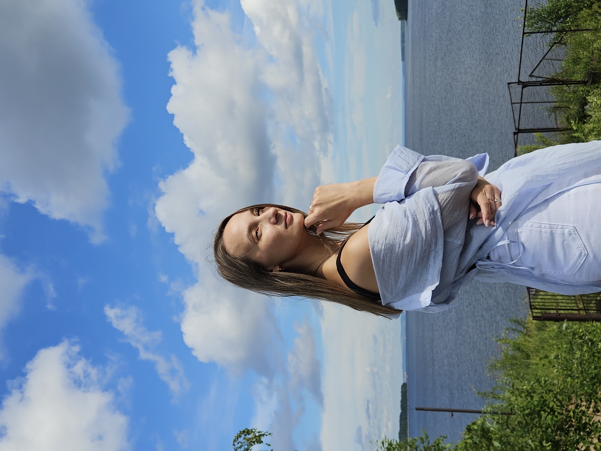 A woman with long, light brown hair, wearing a light blue off-the-shoulder top and white pants, stands on a grassy embankment overlooking a wide river. She is facing to her right, with her chin resting on her hand, and looking out at the water. The sky is a bright blue with fluffy white clouds. In the background, the river stretches out to the horizon, with a tree-lined shore visible in the distance. The location is likely the embankment of the Kama River in the city of Chaikovsky, Russia, given its characteristic wide expanse and the presence of distant shorelines. A metal fence runs along the embankment behind the woman. Green bushes and trees are visible in the foreground.