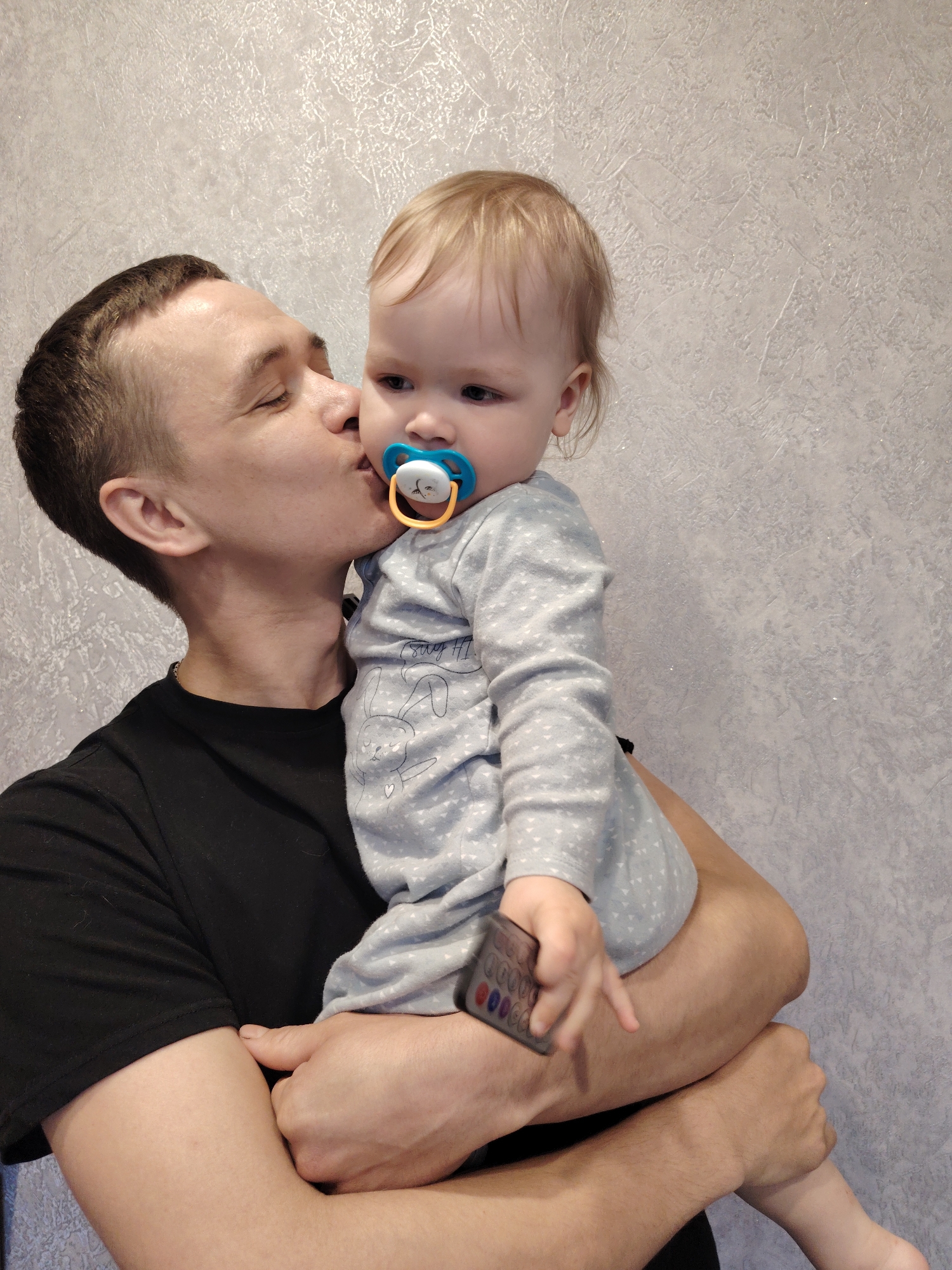 A medium shot shows a man holding a baby. The man is light-skinned with short dark hair, wearing a black t-shirt, and is kissing the baby on the cheek. The baby, who appears to be light-skinned with blonde hair and a pacifier, is wearing a grey patterned long-sleeved shirt and holds a remote control. The background is a textured, light-colored wall. The image was taken in the city of Chaikovsky, Russia.