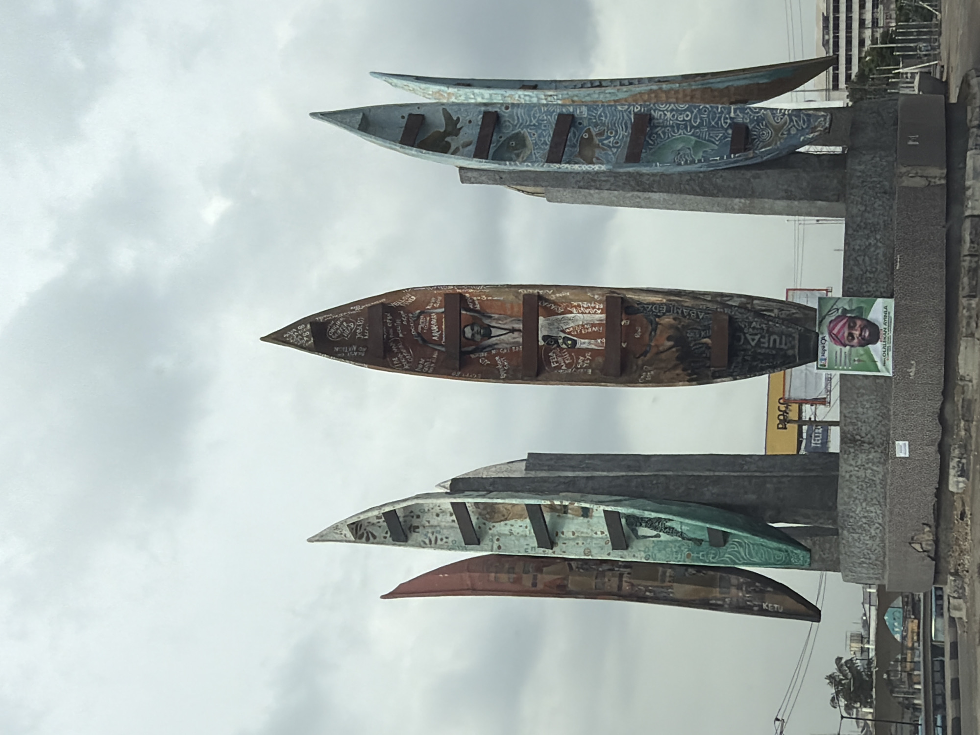 The image shows a sculpture in Ikeja, Nigeria, featuring several dugout canoes arranged vertically on concrete supports. The canoes are painted with various designs, including patterns, figures, and text. A poster is affixed to the base of the sculpture, featuring a man's face. The background includes buildings, signage, and an overcast sky. The sculpture is located on a roadside with urban infrastructure visible.