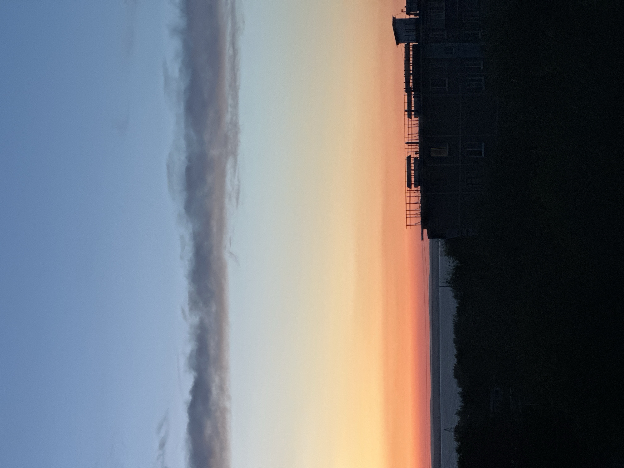 The image shows a sunset over a body of water, with a silhouetted building in the foreground. The sky is a gradient of blue, orange, and pink. The building appears to be a multi-story apartment complex or office building. Trees and other vegetation are visible in the foreground, also silhouetted against the sky. The overall scene is peaceful and serene. There are no people visible in the image. The photograph was taken in the city of Energetik, Russia.
