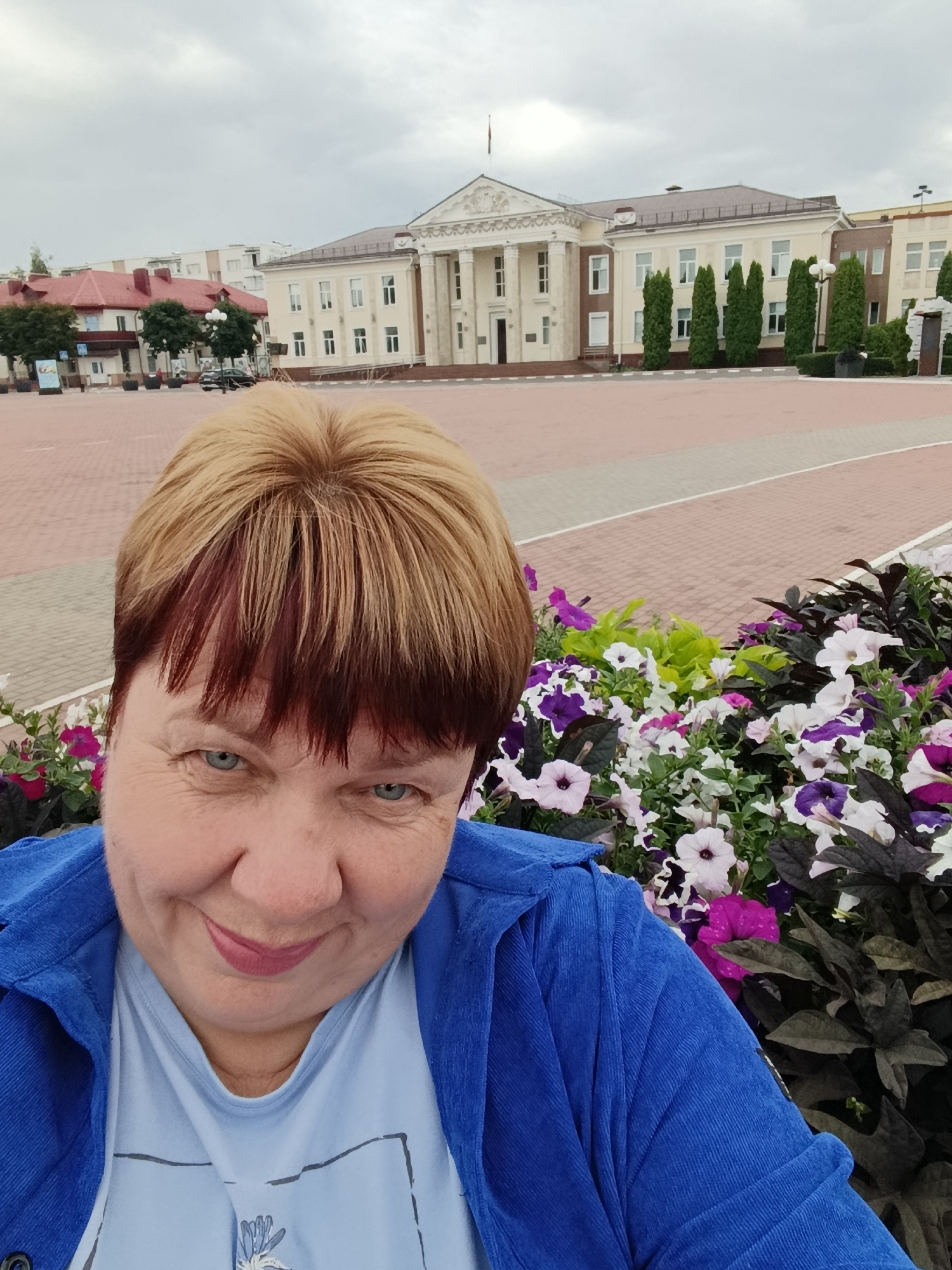 A woman with blonde and auburn hair, wearing a blue jacket and a light blue t-shirt, smiles at the camera. She is in the foreground, with a flowerbed of purple and white petunias behind her. In the background, a large, light-colored building with columns and a flag pole stands under an overcast sky. The building appears to be a government or public institution. The area in front of the building is paved with red and grey interlocking bricks. The scene is consistent with a public square in Zhlobin, Belarus.