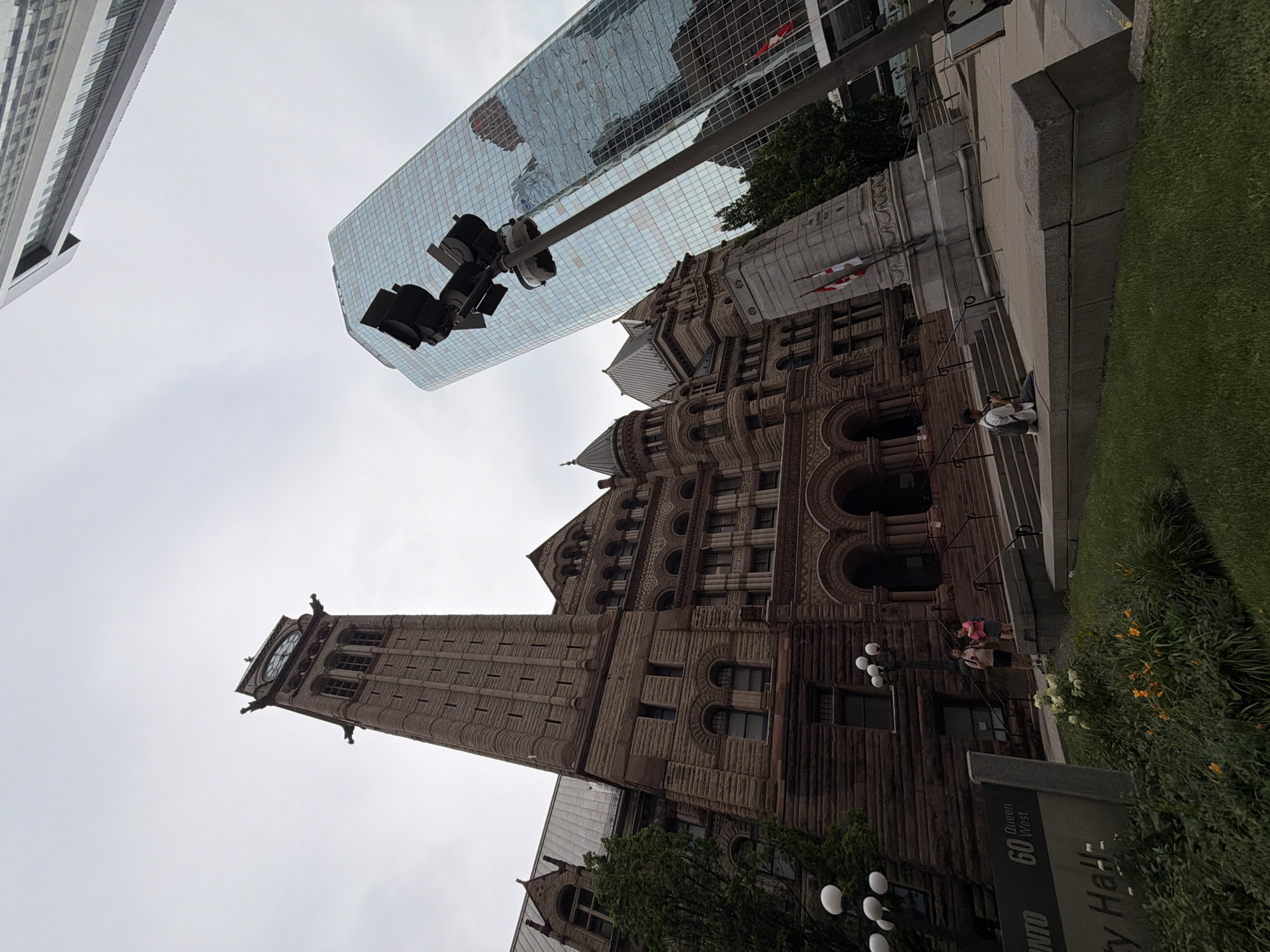The image captures a low-angle view of Toronto's Old City Hall, a historic building with a prominent clock tower, situated next to a modern glass skyscraper. The sky is overcast. In the foreground, a street sign indicates "Toronto 60 Queen West City Hall." People are visible in the scene: a couple stands on the steps of Old City Hall, and a lone individual sits on a stone ledge in the mid-ground, facing away from the camera. A war memorial with Canadian flags is positioned between the two buildings. A traffic light pole stands in the mid-right of the frame, obscuring part of the modern building. The overall impression is an urban landscape showcasing a juxtaposition of historical and contemporary architecture under a muted sky.