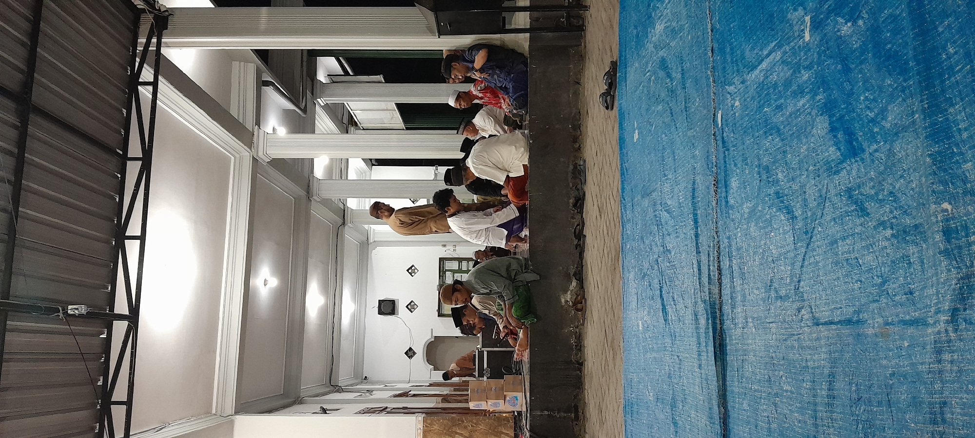 A group of men, predominantly dressed in traditional Islamic attire, are seated and standing on a raised platform in what appears to be an outdoor or semi-covered area. The setting suggests a communal gathering, possibly a religious event or a social meeting in Bendungan, Indonesia. The men are positioned in a relaxed manner, with some engaged in conversation and others appearing contemplative. Several individuals are wearing peci hats, a common headwear in Southeast Asia. Behind the men, a building with white pillars and a corrugated metal roof is visible. In the foreground, a large blue tarp covers the ground, indicating an outdoor space that might be used for various activities or to provide a clean surface for the gathering. The lighting suggests it is either evening or indoors with artificial illumination.