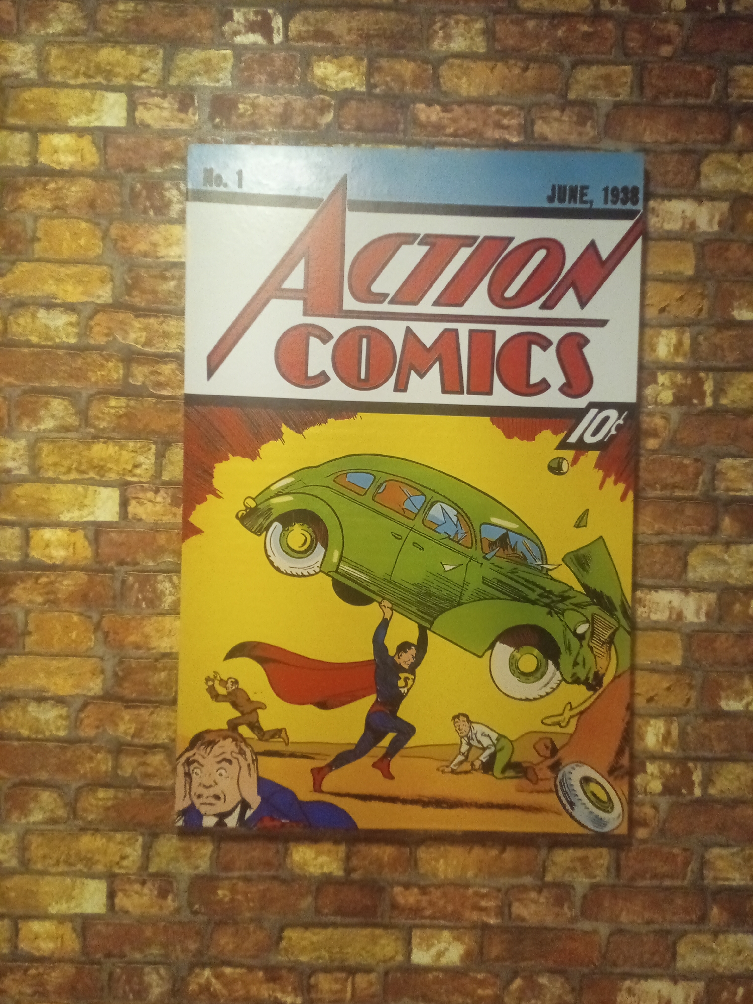 The image shows a framed replica of the cover of Action Comics #1, featuring Superman lifting a car. The replica is mounted on a brick wall. The text on the comic cover reads "Action Comics" and "June, 1938". The comic depicts Superman in his blue suit and red cape, holding up a green car that appears to be crashing. Below, a man in a suit is shown holding his head in distress, while another man in a white shirt and green pants is crawling on the ground. The background is a yellow expanse with red streaks. The brick wall behind the comic has a texture of alternating brown and yellowish-orange bricks. The lighting suggests an indoor setting. This image was taken in Porlamar, Venezuela.