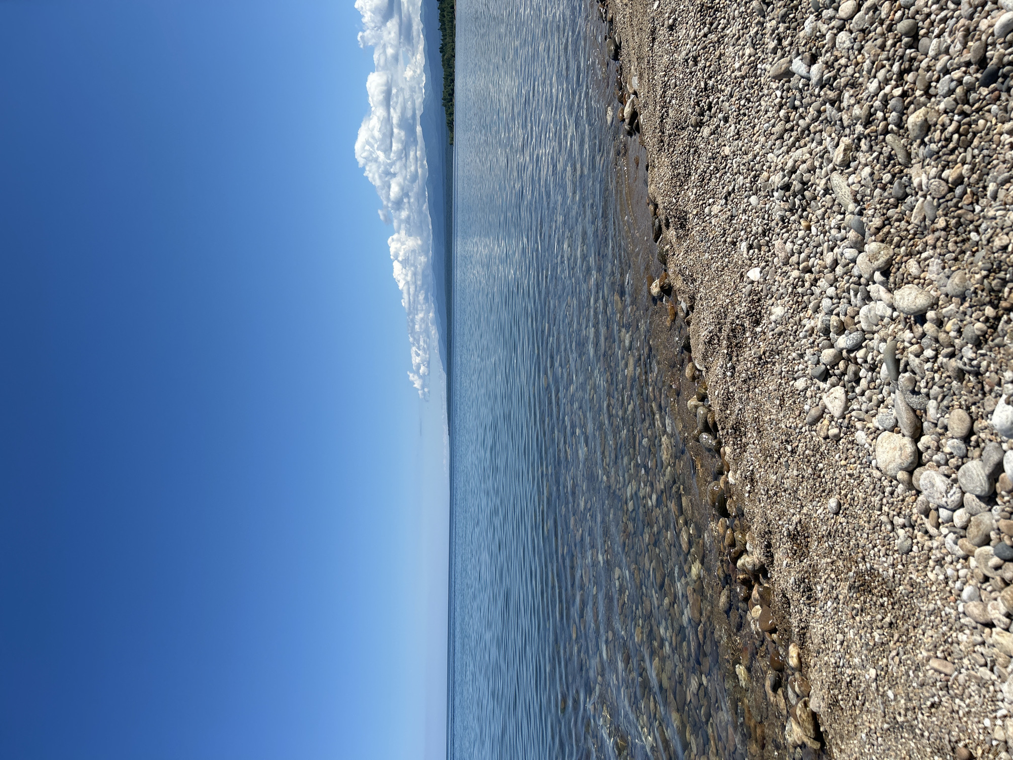 The image shows a wide, tranquil body of water, likely a lake, under a clear blue sky. The water is calm with gentle ripples, and the bottom is visible near the shore, revealing a bed of smooth, rounded pebbles of various sizes and colors. The shoreline is densely covered with these pebbles, creating a natural beach. In the distance, a hazy line of mountains can be seen against the horizon, with some scattered white clouds in the sky. The scene appears to be a natural landscape, devoid of any human presence or artificial structures. Given the context of Tolbazikha, Russia, this is likely a depiction of a shoreline on one of the many lakes in the region.