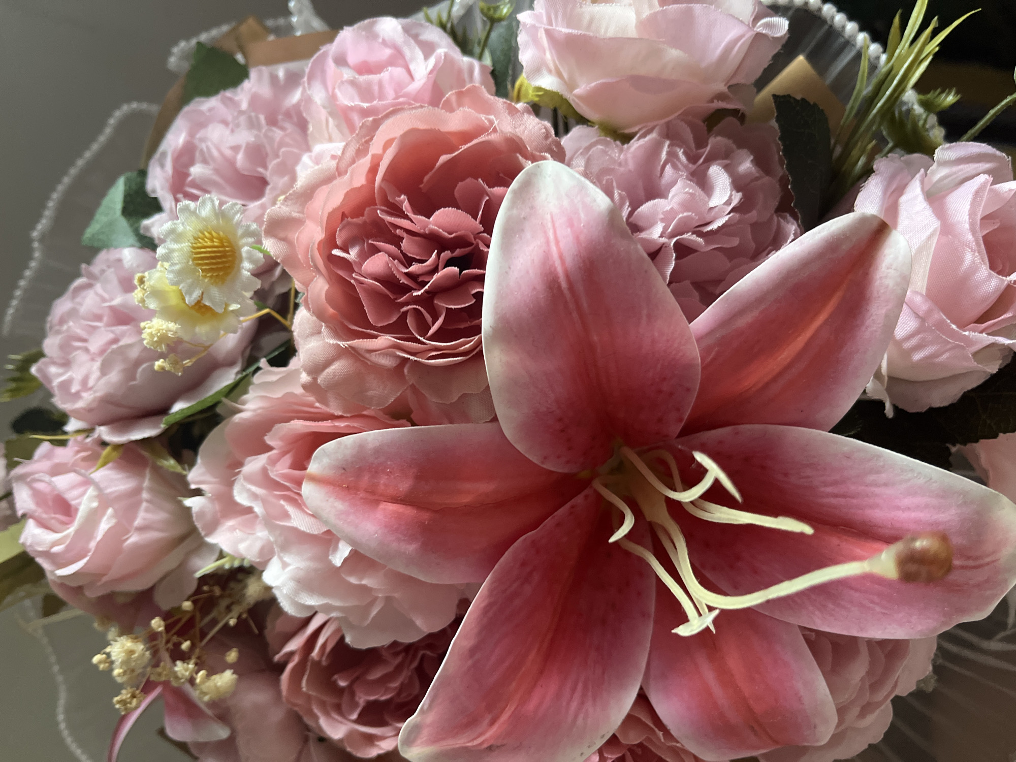 A close-up, eye-level shot focuses on a bouquet of artificial pink flowers, primarily lilies and roses, set against a plain, grey background. The flowers are arranged in a tight cluster, with a large pink lily dominating the foreground. The lily's petals are a vibrant pink with white edges and prominent yellow stamens. Behind the lily, a variety of pink roses and other smaller pink flowers are visible, some with ruffled petals and others with a more delicate appearance. A few small, white, daisy-like flowers with yellow centers are also interspersed among the larger blooms. Green artificial leaves and stems add a touch of contrast to the floral arrangement. The lighting appears to be soft, highlighting the textures and colors of the artificial flowers. There is no indication of a specific location within a city or any people present.