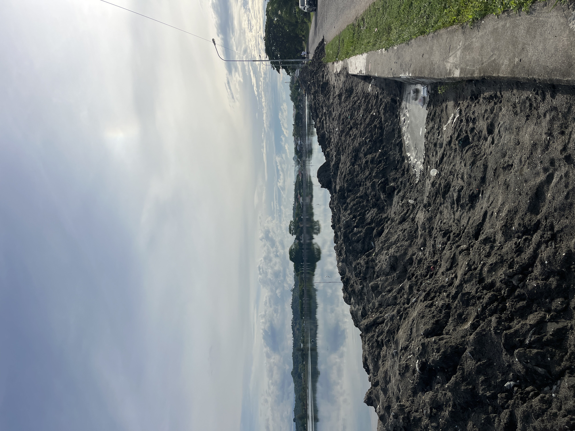 The image depicts a roadside scene in Lhokseumawe, Indonesia. Piles of dark soil or earth line the edge of the pavement, with small puddles visible. The sky is overcast, reflecting in a body of water that mirrors the opposite shoreline. A lamppost stands along the road, and vegetation is visible beyond the soil. A vehicle is parked on the road.