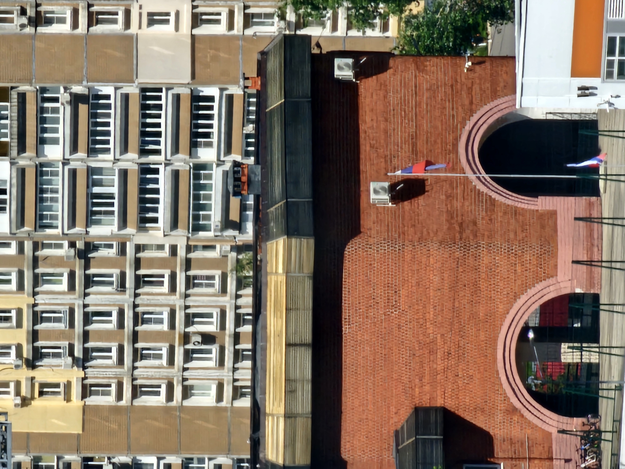This is an aerial, high-angle shot of an urban landscape featuring architectural elements of buildings. The scene is dominated by the textured brickwork of a large building, contrasting with the repetitive, grid-like pattern of windows on adjacent structures.
On the right side of the image, a large, red brick building features a semi-circular architectural detail, possibly an entrance or decorative element. Two air conditioning units are visible on the roof of this brick structure, along with two flags attached to a clothesline. One flag appears to be red, white, and blue, while the other is primarily red. Further to the right, a white and orange building is partially visible.
To the left of the brick building, a narrower section of a building with a dark, possibly metal roof, is present. Adjacent to this is a building with a facade composed of numerous rectangular windows, each divided into several panes by white frames. The windows are set within beige or tan colored panels. Further to the left, another building exhibits a similar window structure, with some windows appearing to be open. The colors of these buildings are primarily beige, tan, and white, with green foliage visible between some of the upper windows.
The overall impression is of a dense urban environment, likely during daylight hours given the shadows cast. The image appears to be a still photograph, with no specific activity or event clearly depicted beyond the presence of the flags and the architectural features. No text is visible in the image. The scene is factual and observational, lacking any overt emotional cues. The location context is Nizhny Novgorod, Russia.