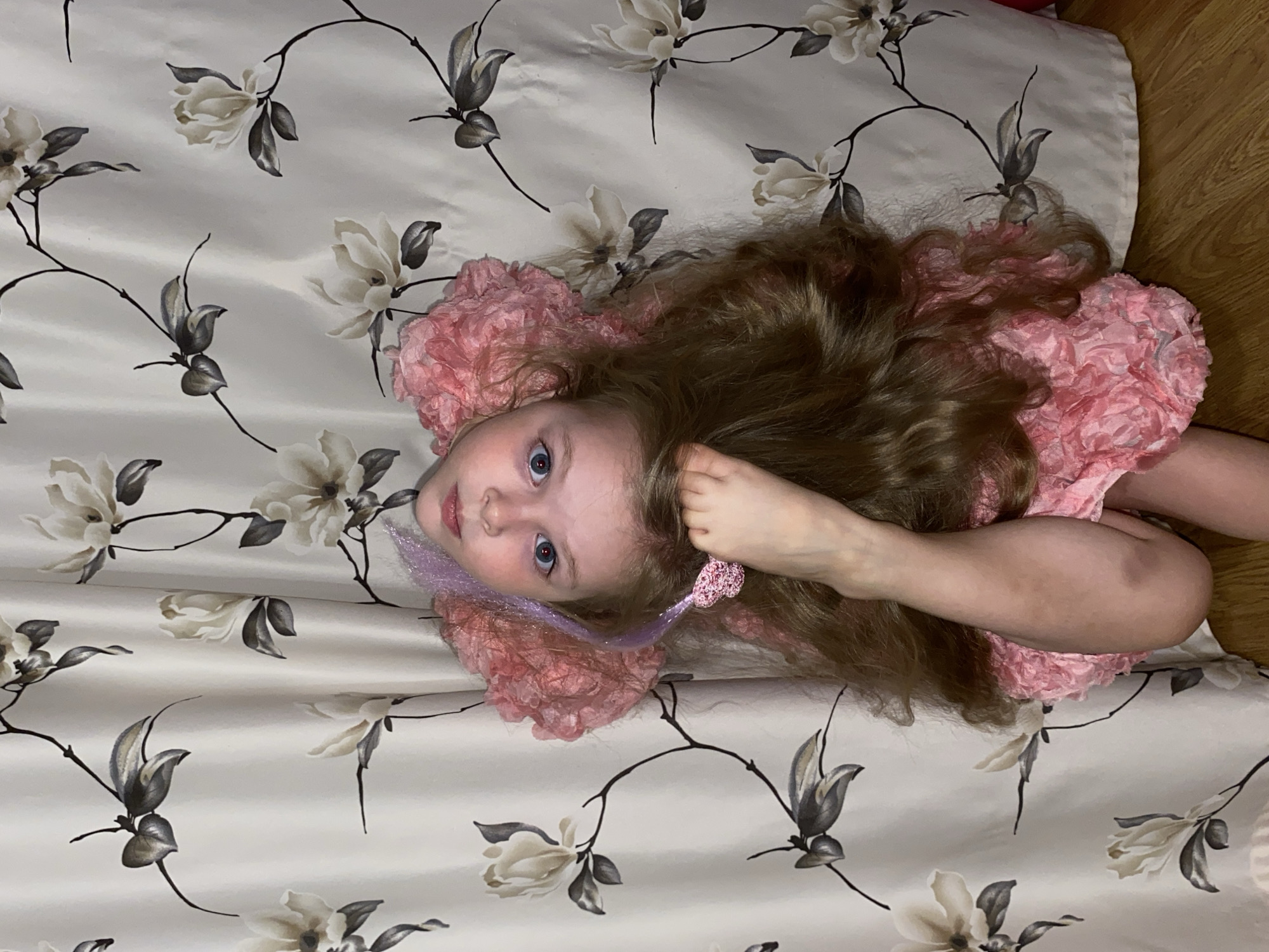 A young girl with long, light brown, wavy hair and striking blue eyes is the central subject of this indoor portrait. She is lying on her back, looking directly up at the camera, with her head slightly tilted. Her expression is neutral and observant. She wears a pink dress with a textured, possibly ruffled or layered, fabric. A light purple, sparkly hair extension or accessory is visible near her left cheek, and a small pink, sparkly heart-shaped clip is in her hair near her right temple. Her left arm is bent, with her hand resting near her head.
The setting appears to be indoors, with the girl positioned against a light-colored fabric that serves as the background. This fabric features a recurring pattern of magnolia-like flowers in shades of off-white and grey, with dark, slender stems and leaves. The fabric shows subtle creases, suggesting it might be bedding, a duvet cover, or a large piece of cloth. A small section of warm-toned wooden flooring or furniture is visible along the right edge of the image. The lighting is soft and even, indicating an indoor environment where the time of day is not distinctly marked. No specific activity beyond posing for the photograph is depicted. There is no visible text in the image. The location context provided is Votkinsk, Russia.