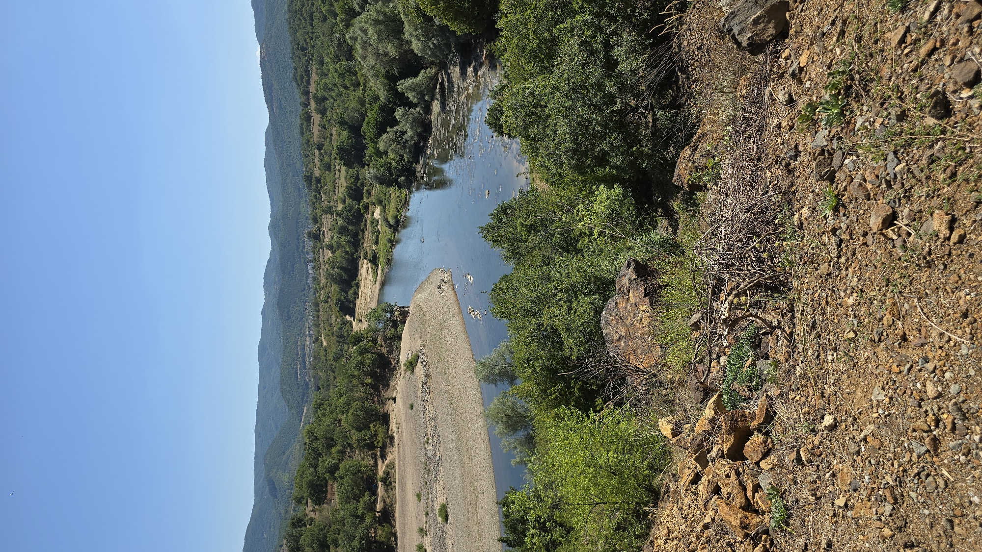 The image depicts a wide, winding river flowing through a valley with rolling, tree-covered hills in the background. The foreground is a barren, rocky slope with sparse vegetation and dry brush. The river has sandy banks in some areas and is bordered by dense green trees. The water appears calm, with some debris floating on the surface in the middle ground. In the distance, there is a small town visible nestled amongst the hills. The sky is clear and blue. The overall impression is a natural landscape under a bright, sunny sky, likely during daytime. The scene is consistent with a view overlooking the Arda River near the town of Madzharovo, Bulgaria, known for its scenic river valleys and hilly terrain. There are no people or specific actions evident in the image.