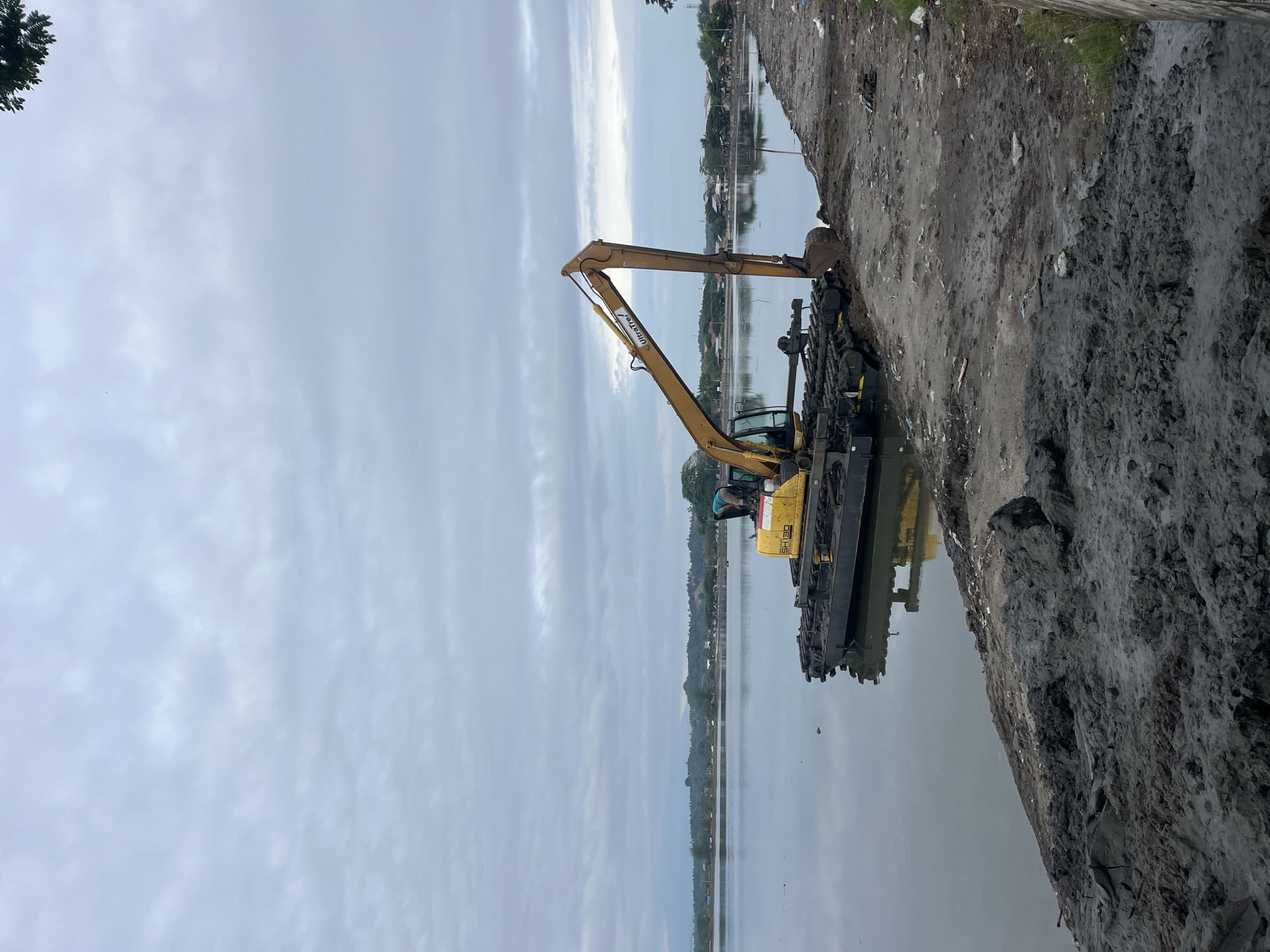 The scene depicts a yellow excavator positioned on a muddy bank next to a body of water, likely in Lhokseumawe, Indonesia. The excavator's arm is extended, suggesting it is in operation. The surrounding environment includes a waterline reflecting the sky, and a muddy shoreline littered with debris. The sky is overcast. A few houses are visible in the distance on the other side of the water.