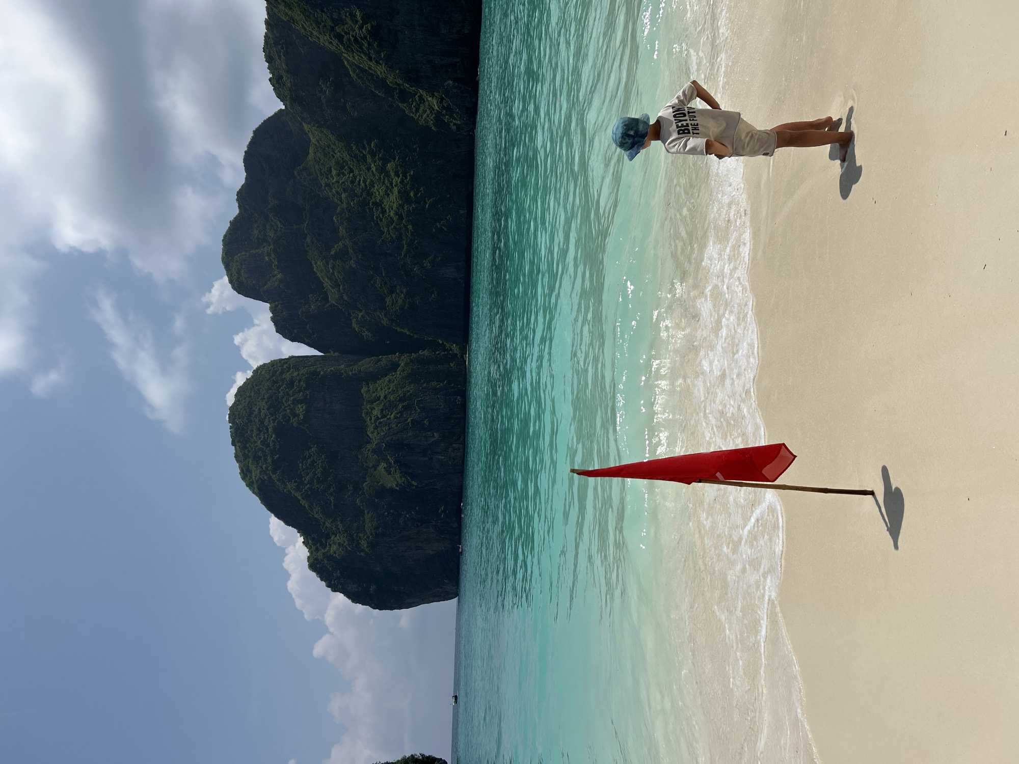 The image depicts a tropical beach scene, likely in Ao Nang, Thailand, characterized by a bright sandy shore, clear turquoise waters, and lush green karst cliffs in the background. In the foreground, a young child wearing a blue bucket hat and a light-colored t-shirt with text visible on the back stands facing the water, with their hands on their hips. A red flag on a wooden pole is planted in the sand to the left of the child, casting a shadow. The water has gentle waves lapping the shore, and several small boats are visible in the distance. The sky is blue with scattered white clouds.