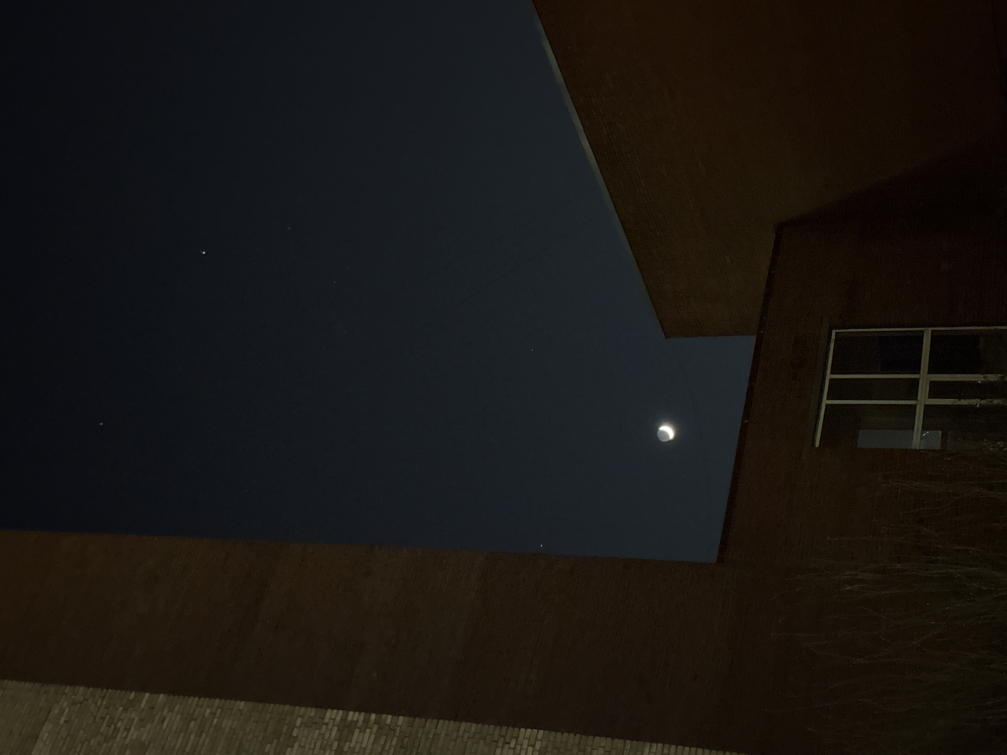 A low-angle, nighttime photograph shows a view of the sky framed by the edges of buildings. A crescent moon is visible in the upper-right quadrant of the sky. Several smaller, dimmer points of light, possibly stars, are also visible. A window with a grid-like pattern is seen on the right side of the frame. The walls of the buildings are dark brown, with a brick-like texture visible on one side. The location is likely Saint Petersburg, Russia, given the urban architecture and the time of year when a crescent moon might be visible in the night sky.