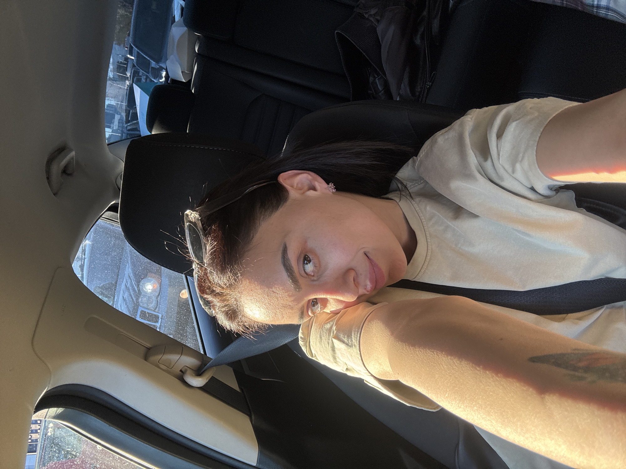 An individual is captured in a car interior, positioned sideways relative to the camera, with strong, warm sunlight illuminating their face and right arm. The person has dark hair, wears a light-colored t-shirt, and has sunglasses resting on top of their head. A small earring is visible on their left ear, and a tattoo can be seen on their right forearm. They are leaning into the shot, resting their head on their right hand, which is propped against their arm, creating a relaxed pose.
The car's interior features black seats and a visible seatbelt. Through the windows, glimpses of an urban environment are visible, including multiple buildings and what appears to be a large vehicle with a distinct circular light. The intense sunlight indicates it is daytime, likely morning or late afternoon, under clear weather conditions. The person maintains a relaxed, neutral expression, with a slight hint of a smile, directly engaging with the camera. There is no visible text in the image. The setting is Tbilisi, Georgia.