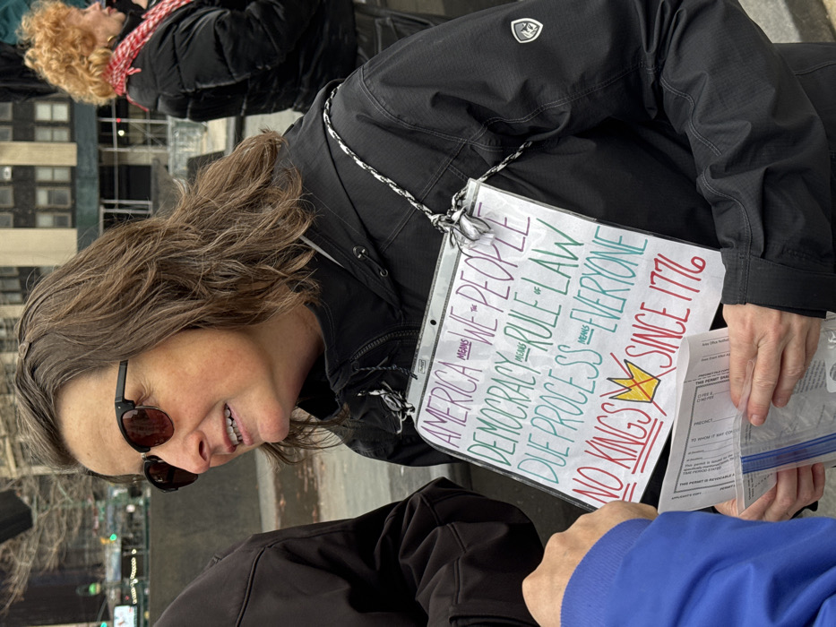 A woman wears sunglasses and a black jacket with a logo on the upper left. She holds a sign that reads "AMERICA WE THE PEOPLE, DEMOCRACY RULE OF LAW, DUE PROCESS EVERYONE, NO KINGS SINCE 1776," accompanied by a drawing of a crown. She also holds what appears to be a permit application. Another person in a blue shirt stands next to her. A blurred figure with red hair is visible in the background.