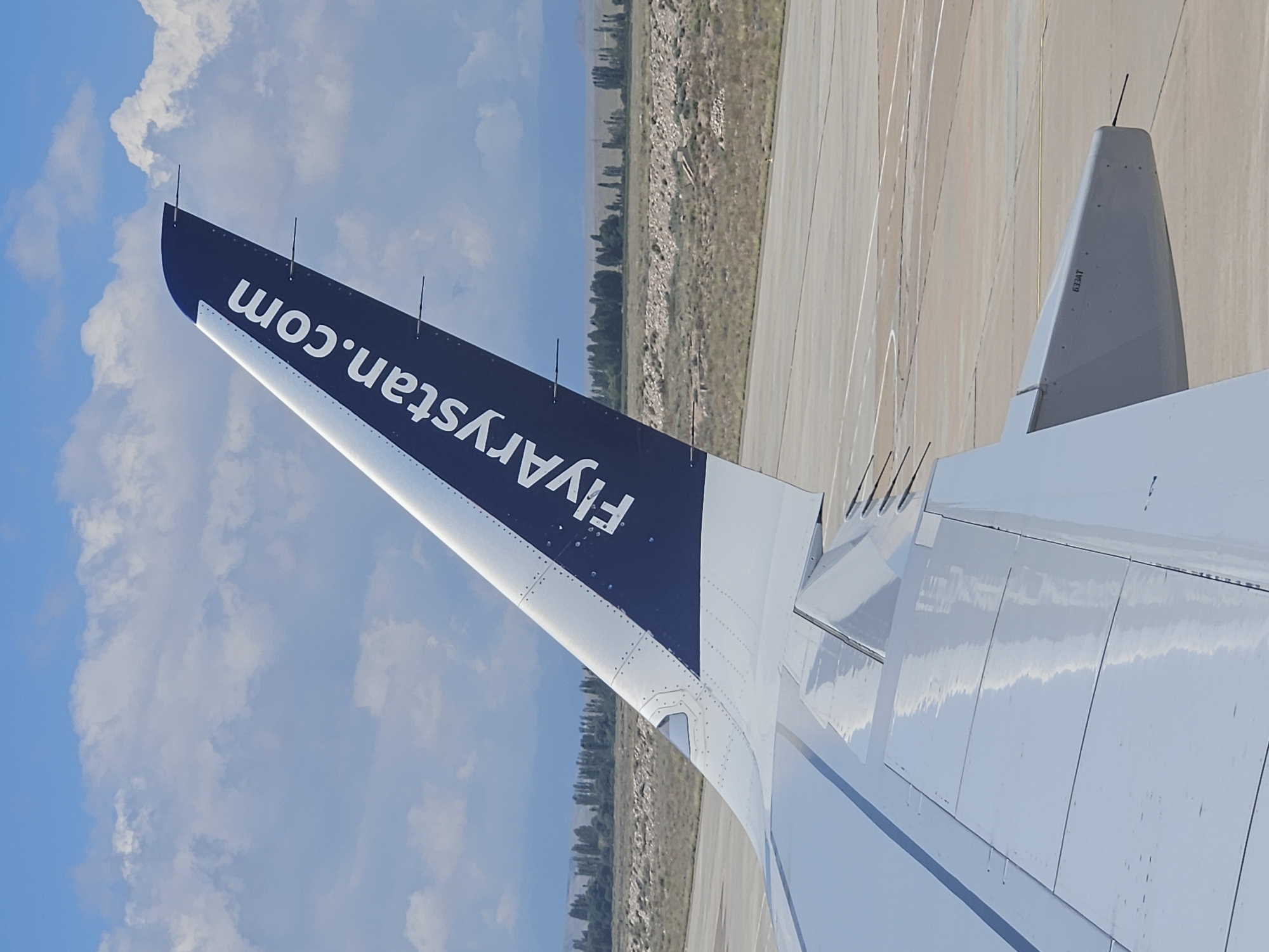 The image shows the tail fin and a portion of the wing of an airplane, with the text "FlyArystan.com" visible on the tail fin. The airplane is on an airport tarmac, and the background shows a dry, arid landscape with some trees and hills, under a cloudy blue sky. The scene appears to be a daytime airport environment. The location is identified as the city of Choktal, Kyrgyzstan.