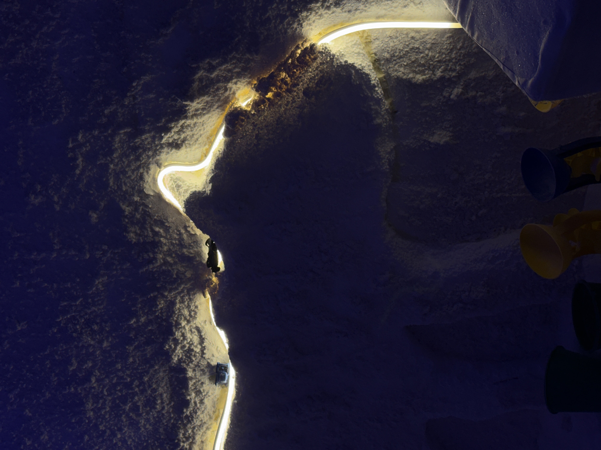 The image depicts an interior space, possibly a cave or tunnel, illuminated by a meandering neon light. The walls appear to be covered in textured material, possibly snow or ice. Two small figures are visible on the left side of the image along the illuminated path. On the right side of the image are three colored megaphones. Without any additional context or identifiers I am unable to confirm the location.