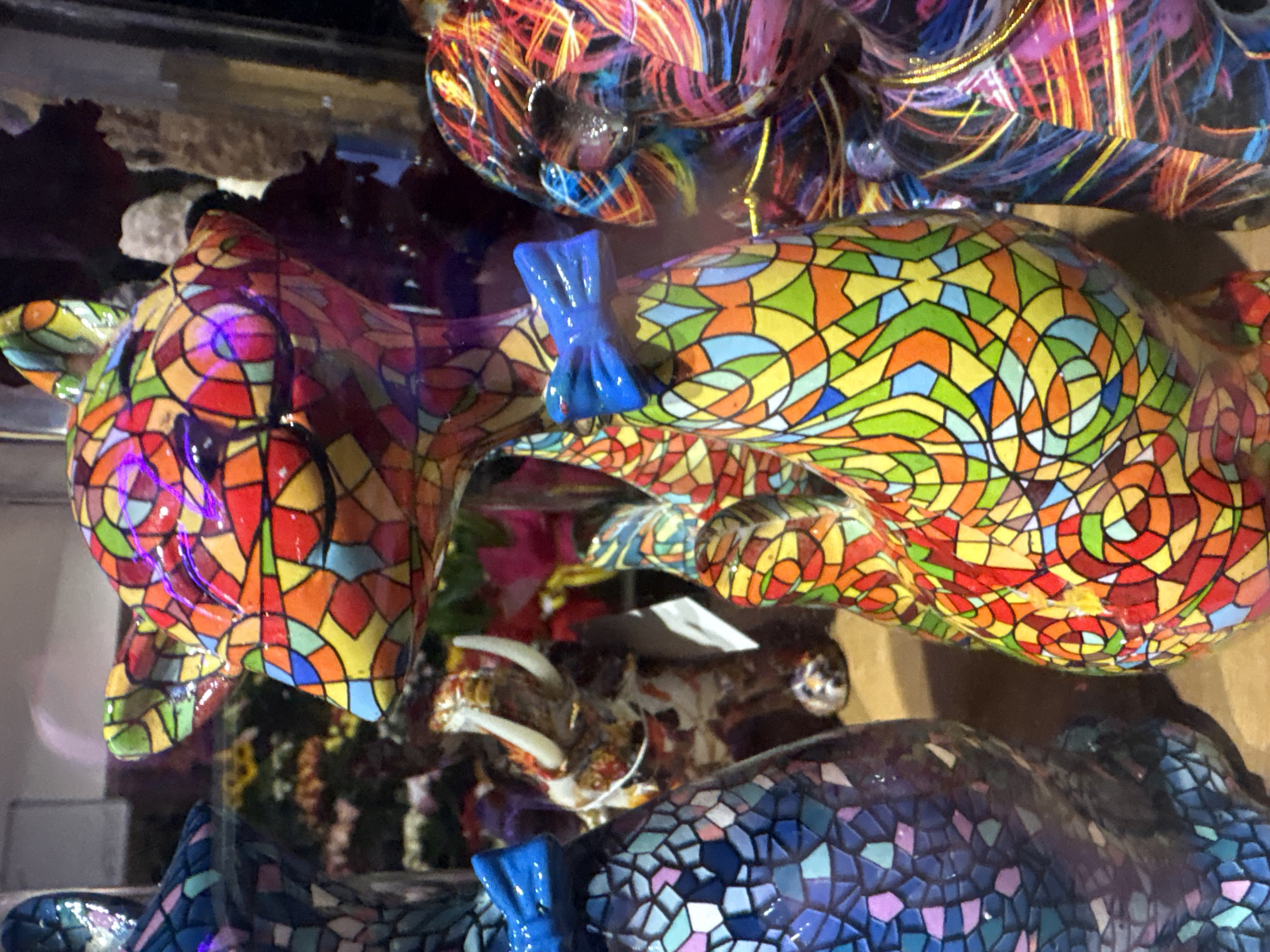 The image displays a retail display featuring multiple ceramic animal figurines, predominantly rendered in a mosaic or stained-glass pattern, behind a reflective surface, likely a glass barrier. The scene is situated within a retail establishment in Sofia, Bulgaria, likely a gift shop, souvenir store, or a dedicated section within a department store or shopping mall, commonly found in commercial districts such as Vitosha Boulevard or major shopping centers.
In the foreground, slightly off-center to the right, is a prominent statue of a feline figure, resembling a cat, sitting upright with its eyes closed. This figure is intricately patterned with irregular, tessellated shapes in vibrant colors including red, orange, yellow, green, blue, and purple, outlined in black, resembling a mosaic or stained-glass effect. A bright blue bow tie is affixed to its neck.
To the left, partially visible, is another similar feline statue, also sitting upright and adorned with a mosaic pattern, but predominantly in shades of blue, teal, and purple. A matching bright blue bow tie is visible on its neck. Only its lower torso and head are clearly within the frame.
Further back in the central area, positioned between the two foreground felines, a smaller ceramic elephant figurine with prominent white tusks is visible. It features a mottled, multi-colored pattern, distinct from the mosaic style of the felines.
On the far right, a portion of a third large ceramic figurine is visible, characterized by an abstract pattern of swirling, multi-directional lines in bright colors (blue, red, yellow, pink) against a dark background, also suggesting a glossy finish.
The figurines rest on a light brown, flat surface. The background is out of focus, revealing hints of other merchandise or display elements, including blurred floral patterns in shades of yellow, green, and red, consistent with a retail environment. Reflections are visible across the glossy surfaces of the figurines and the clear barrier separating them from the viewer, indicating artificial illumination within the display area. No people are visible in the frame.