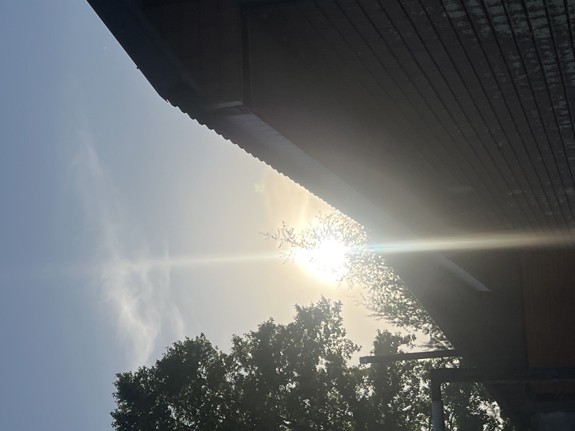 The image appears to be taken from a low angle, looking upwards towards the sky, a building's roof, and trees. The sun is visible, obscured somewhat by tree branches, causing a lens flare effect. The sky is a pale blue with some clouds. The roof overhang dominates the upper part of the image. The lower portion features tree foliage.
I am unable to confirm the location to be Chaikovsky, Russia.