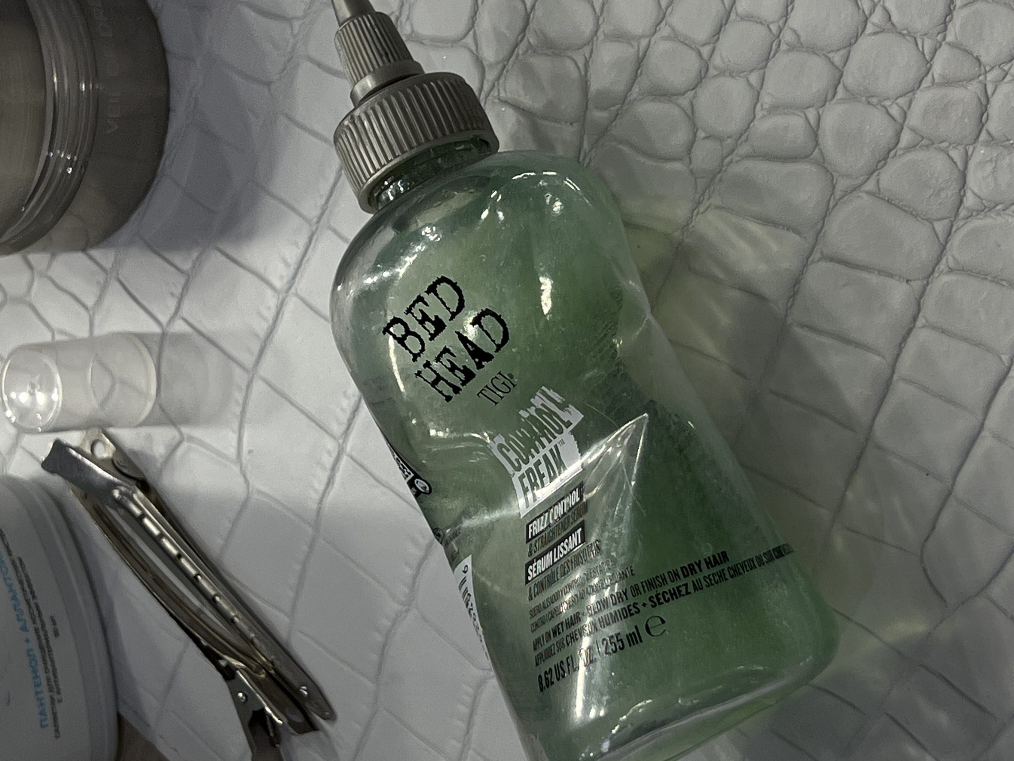 The image presents a close-up, overhead view of hair care products and styling tools arranged on a textured, light grey surface resembling crocodile skin.
The central focus is a transparent, light green bottle of "BED HEAD TIGI CONTROL FREAK" hair product, filled with a gel-like substance and topped with a grey, ribbed dispenser nozzle. The label on the bottle clearly identifies it as a "FRIZZ CONTROL & STRAIGHTENER SERUM" (SÉRUM LISSANT), with instructions to "APPLY ON WET HAIR + BLOW DRY OR FINISH ON DRY HAIR" ("APPLIQUER SUR CHEVEUX HUMIDES + SÉCHEZ AU SÈCHE CHEVEUX OU SUR CHEVEUX"). The volume is marked as "8.62 US FL OZ / 255 ml e."
To the upper left, a partial view of a silver, round container is visible, bearing the text "VEIL OF DREA." Below this, a section of a white container shows Cyrillic text, including "ПАНТЕНОЛ • АЛЛАНТ." A small, clear plastic cap or container is situated near the center-left of the frame. Further to the left, two metallic hair clips, possibly duckbill or alligator clips, are laid on the textured surface.
The scene is illuminated by artificial light, casting soft shadows from the objects. No people are present, and no specific activity is occurring, but the arrangement implies preparation for hair styling or personal grooming.