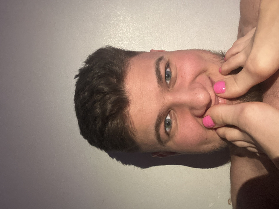 The image displays a close-up frontal view of a young adult male against a plain, light-colored wall, presumed to be indoors in Corby, United Kingdom. The male has short, dark brown hair, blue eyes, and light stubble on his cheeks and chin. He is smiling broadly, looking directly at the camera. Two bare feet, featuring bright pink polished toenails, are positioned directly over his lower face. Specifically, the big toe of the left foot presses against his upper lip and nose area, while the toes of the right foot are situated near his lower lip and chin. Portions of his bare shoulders are visible, suggesting he is shirtless. Lighting appears to originate from the front-left, casting a clear shadow of the subject's head onto the wall behind him. The interaction between the feet and the subject's face conveys a playful or intimate pose.