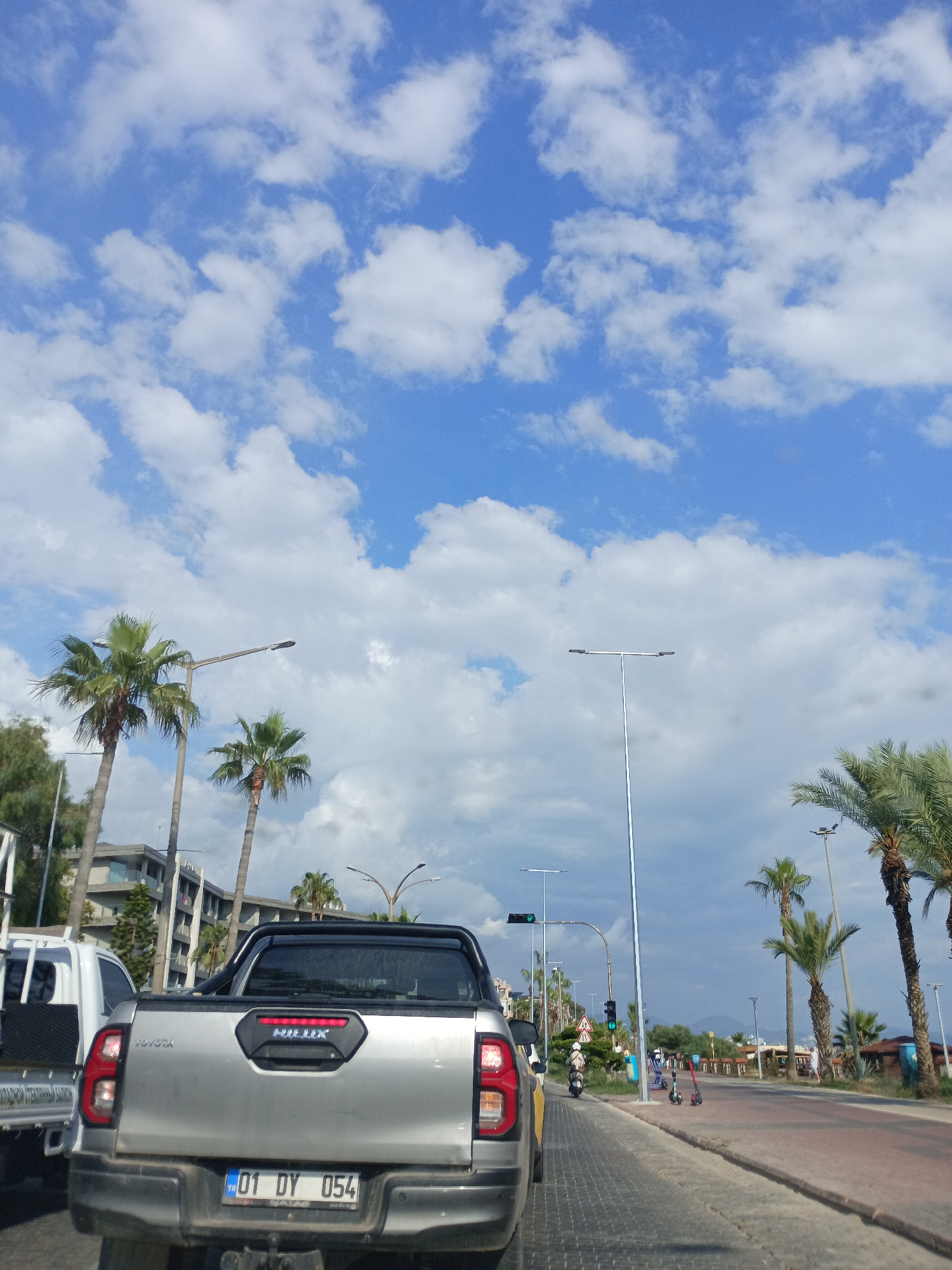 The image captures a bright, sunny day in Alanya, Turkey, with a vibrant blue sky filled with fluffy white clouds. The scene appears to be a street, likely a coastal road given the presence of palm trees lining the pathway.
In the foreground, a silver Toyota Hilux pickup truck is prominently displayed, seen from the rear. Its license plate reads "01 DY 054". Beside it, a white utility vehicle with a black cargo area is partially visible.
Further down the road, several palm trees stand tall, some with their fronds reaching towards the sky. Streetlights are scattered along the road, one of which is a tall, modern design with two lamps. Traffic signals are visible, with one displaying a green light.
The road itself is paved with red bricks and grey asphalt. A few individuals are seen on the sidewalk and the road. One person is riding a scooter, while two electric scooters are parked. In the distance, buildings suggestive of hotels or apartments are visible, interspersed with more greenery. The overall atmosphere seems relaxed and pleasant, typical of a holiday destination. The time of day is likely midday or early afternoon, judging by the bright sunlight and clear sky.