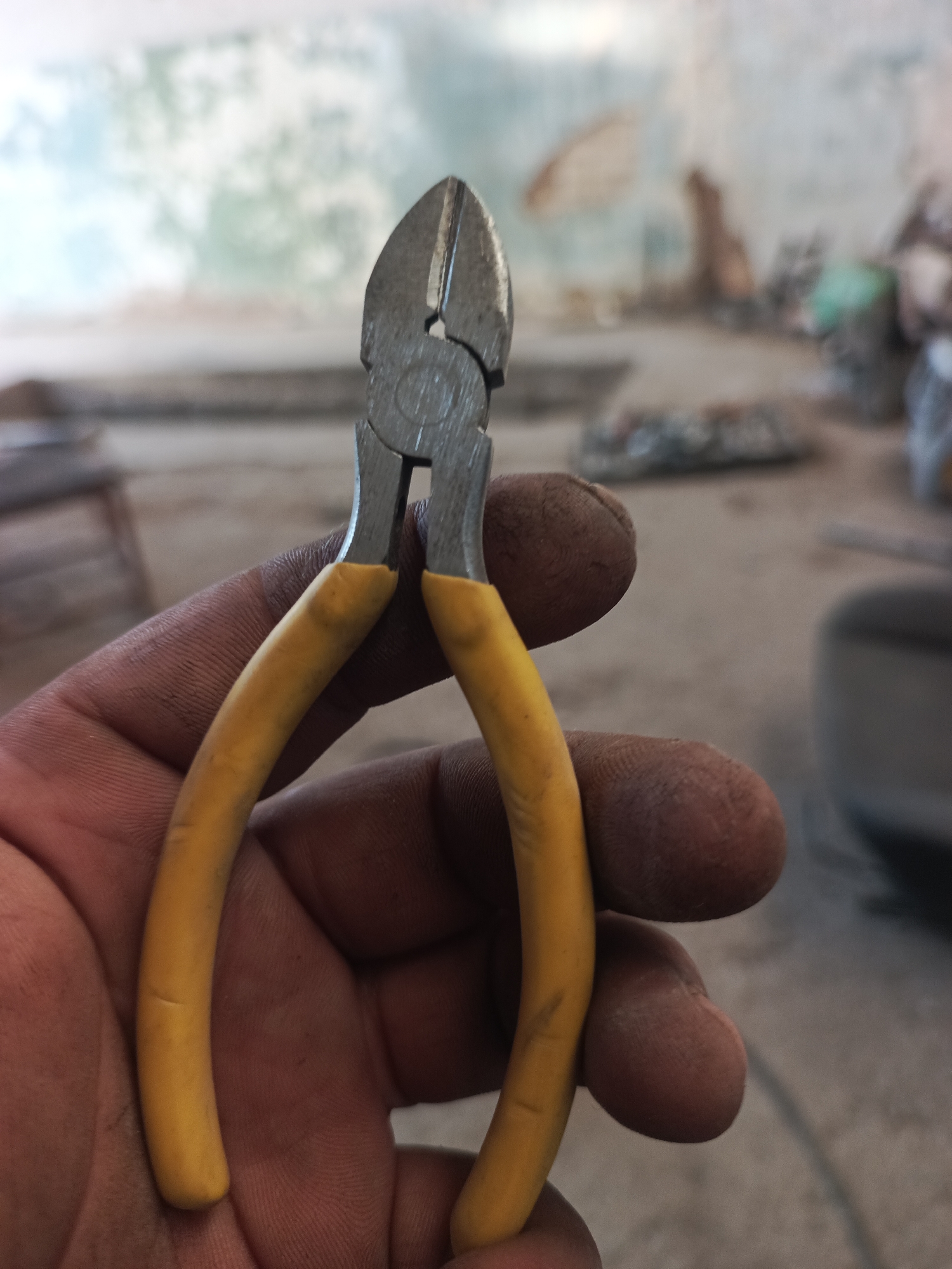 A close-up, over-the-shoulder shot shows a person's hand holding a pair of yellow-handled diagonal cutters. The cutters are made of metal and are positioned in the center of the frame, with their jaws open. The hand is dusty, indicating manual labor. The background is blurred and appears to be an outdoor, possibly construction or workshop environment, with a wall in the distance showing signs of wear and tear. The scene is set in Kumkurgan, Uzbekistan.