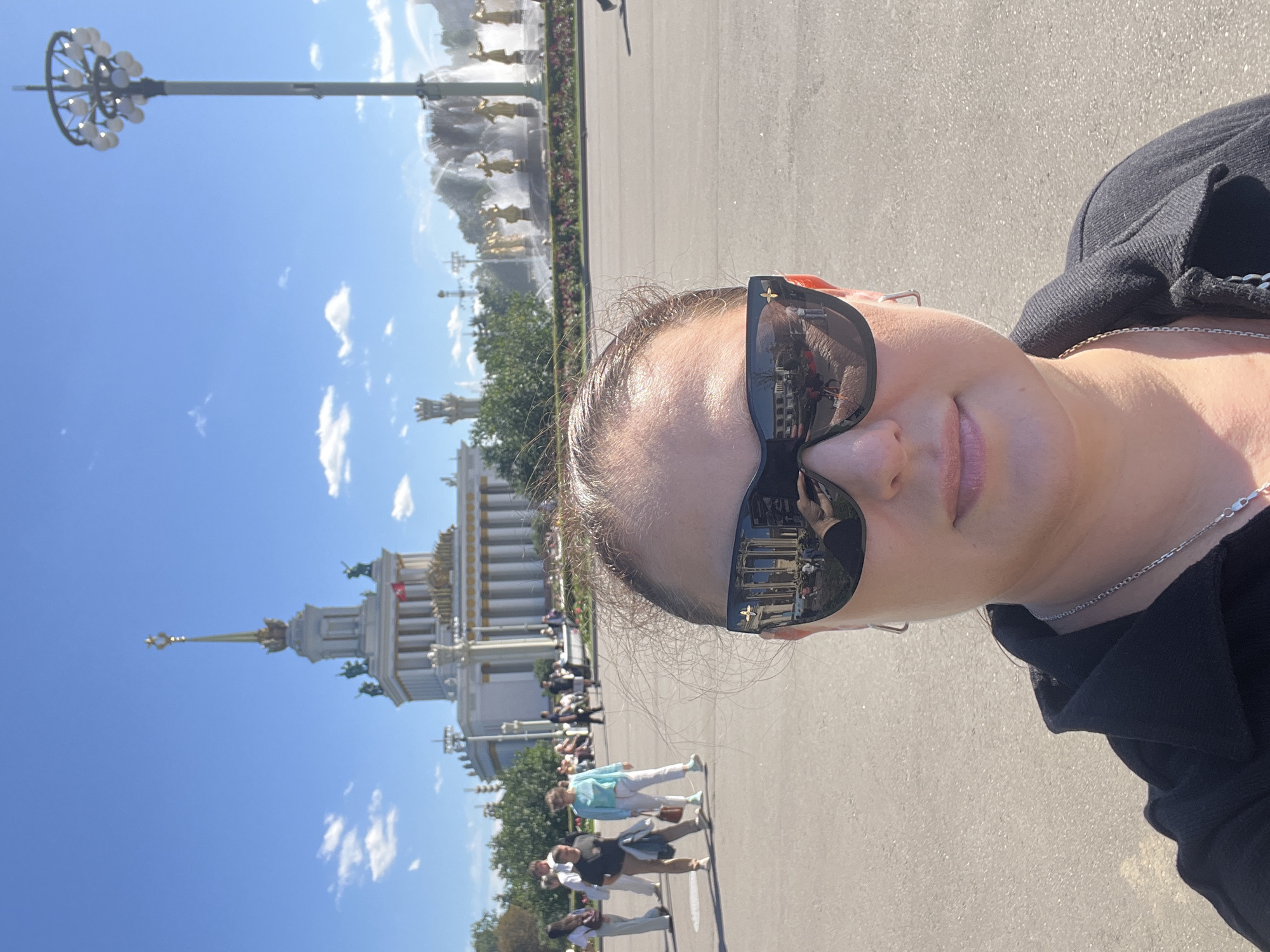 A woman takes a selfie in front of a large, ornate building with a tall spire. She is wearing sunglasses and a black jacket. In the background, a fountain with golden statues is visible, as well as several other people walking around. The scene appears to be a public square in Moscow, Russia, likely VDNKh, given the architecture. The sky is blue with scattered clouds, and the sun is shining.