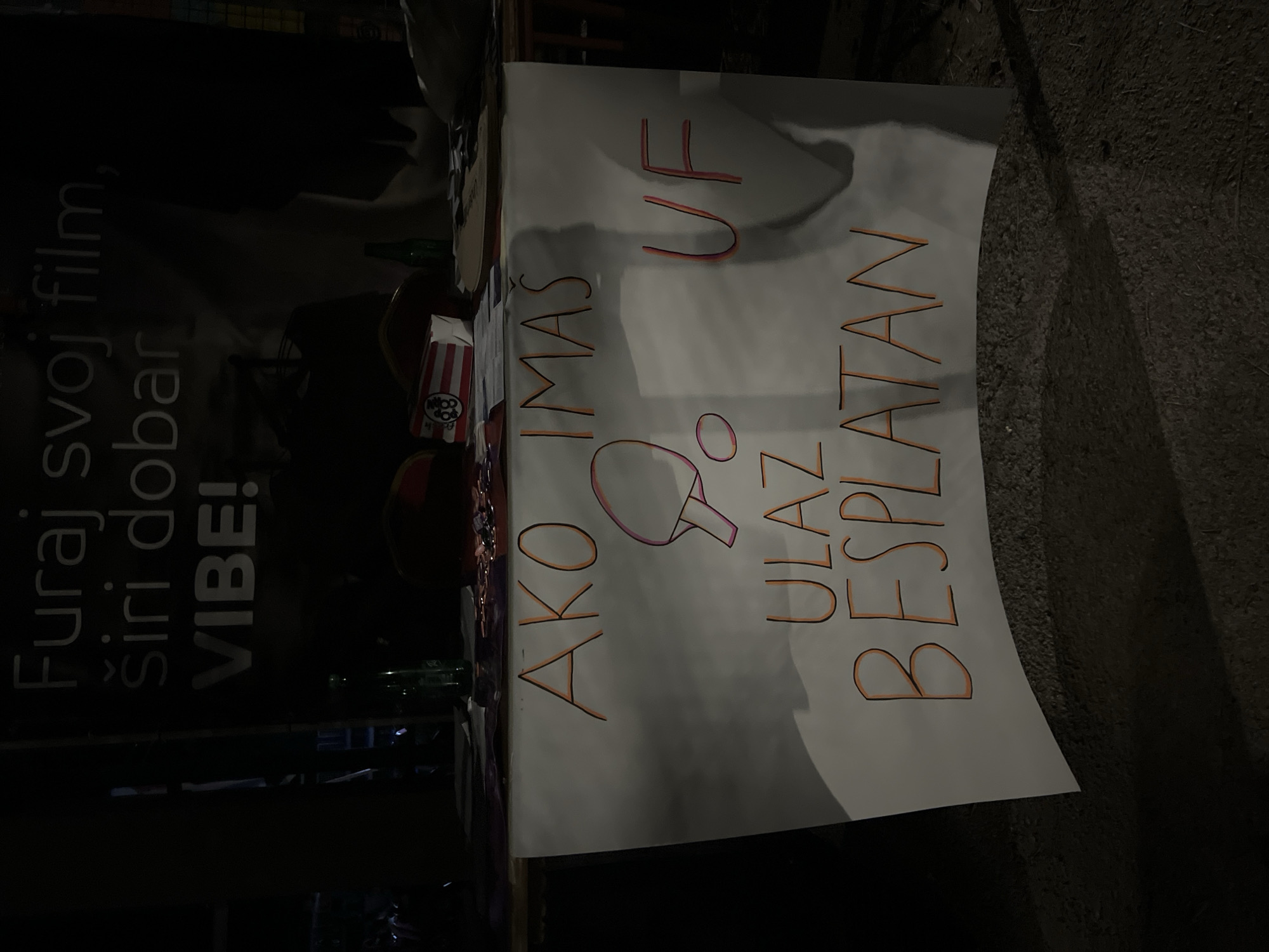 The image captures a nighttime scene in Podgorica, Montenegro, focusing on a table with a large white sheet of paper on which text is written in orange and pink marker. The text appears to be in Serbian or a similar Slavic language. The overall message seems to be promotional or informational, with phrases like "Furaj svoj film, širi dobar VIBE!" ("Play your movie, spread good VIBE!") visible on a banner in the background, and "AKO IMAS", "UF", "ULAZ BESPLATAN" ("IF YOU HAVE", "UF", "ENTRANCE FREE") on the foreground sheet. A drawing of a ping pong paddle and ball is also present. On the table, there are two green bottles, a red and white popcorn container with a logo, and other assorted items. The lighting is dim, suggesting an outdoor event or market setting at night, with shadows cast on the ground and the banner.